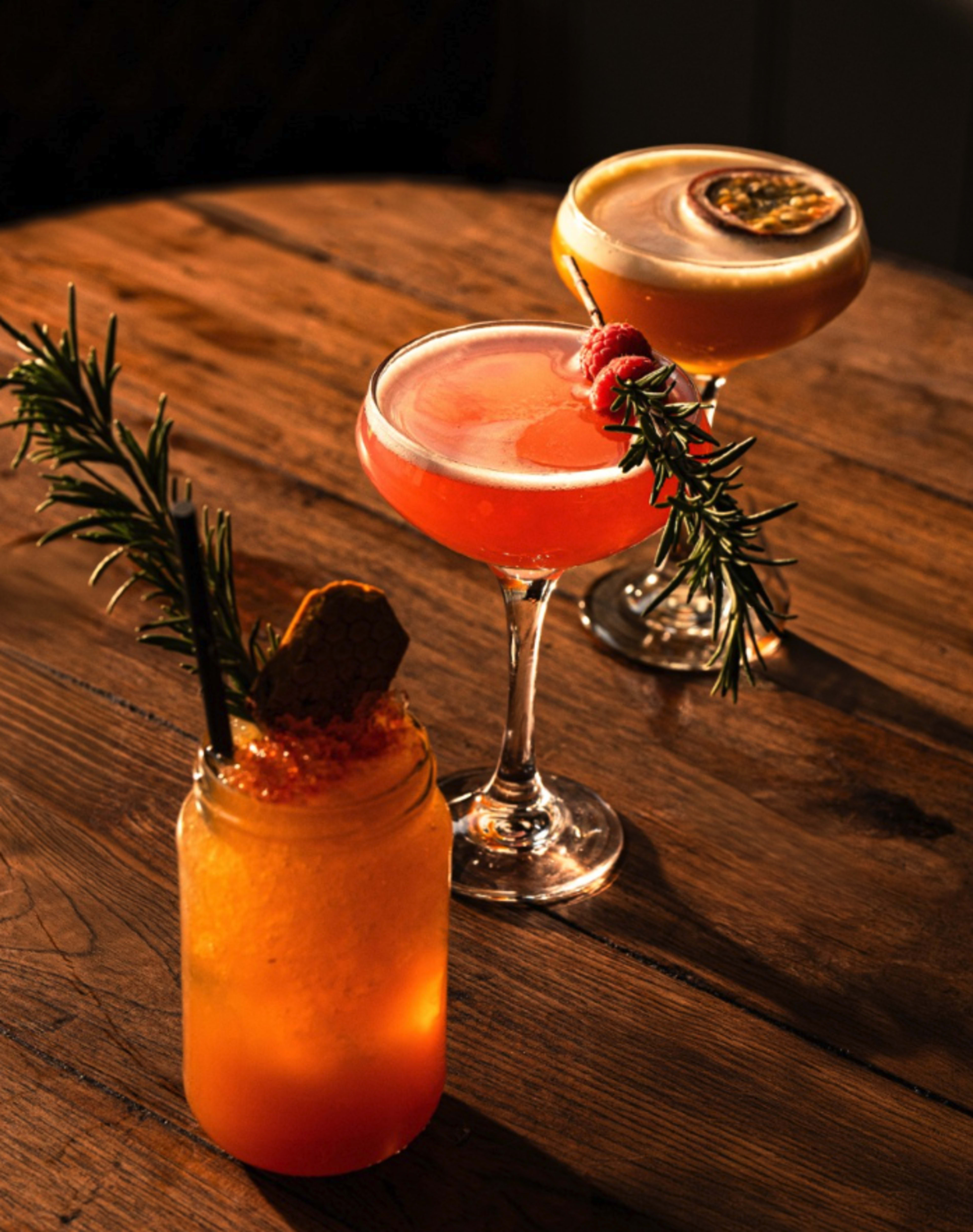 Three colorful cocktails are arranged on a wooden table, with one garnished with a sprig of rosemary and a honeycomb piece, while the others are served in elegant glasses.