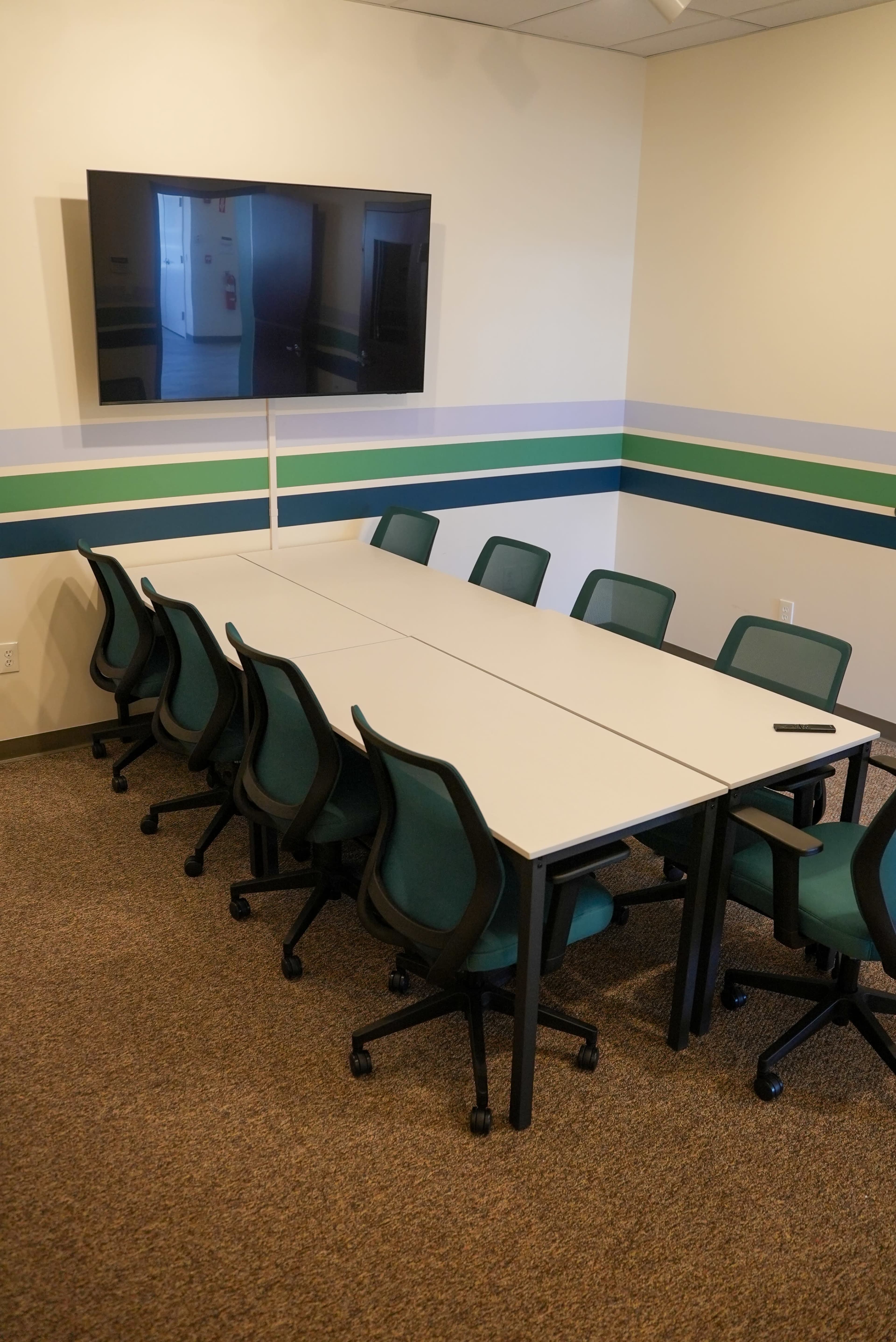 A rectangular meeting table with a row of chairs is positioned in a conference room featuring a wall-mounted television.