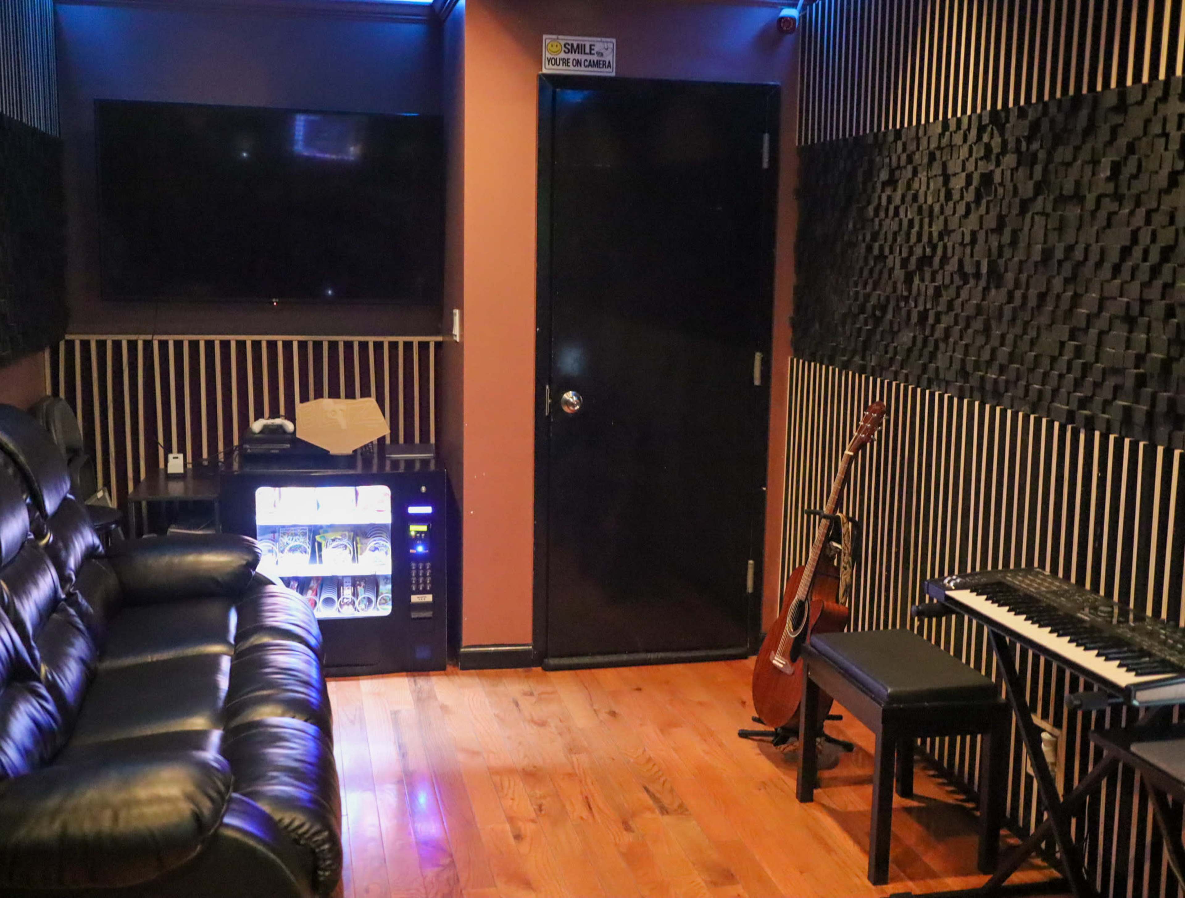 A small recording studio with a black leather couch, a small beverage fridge, a closed door, and musical equipment including a keyboard and an acoustic guitar.