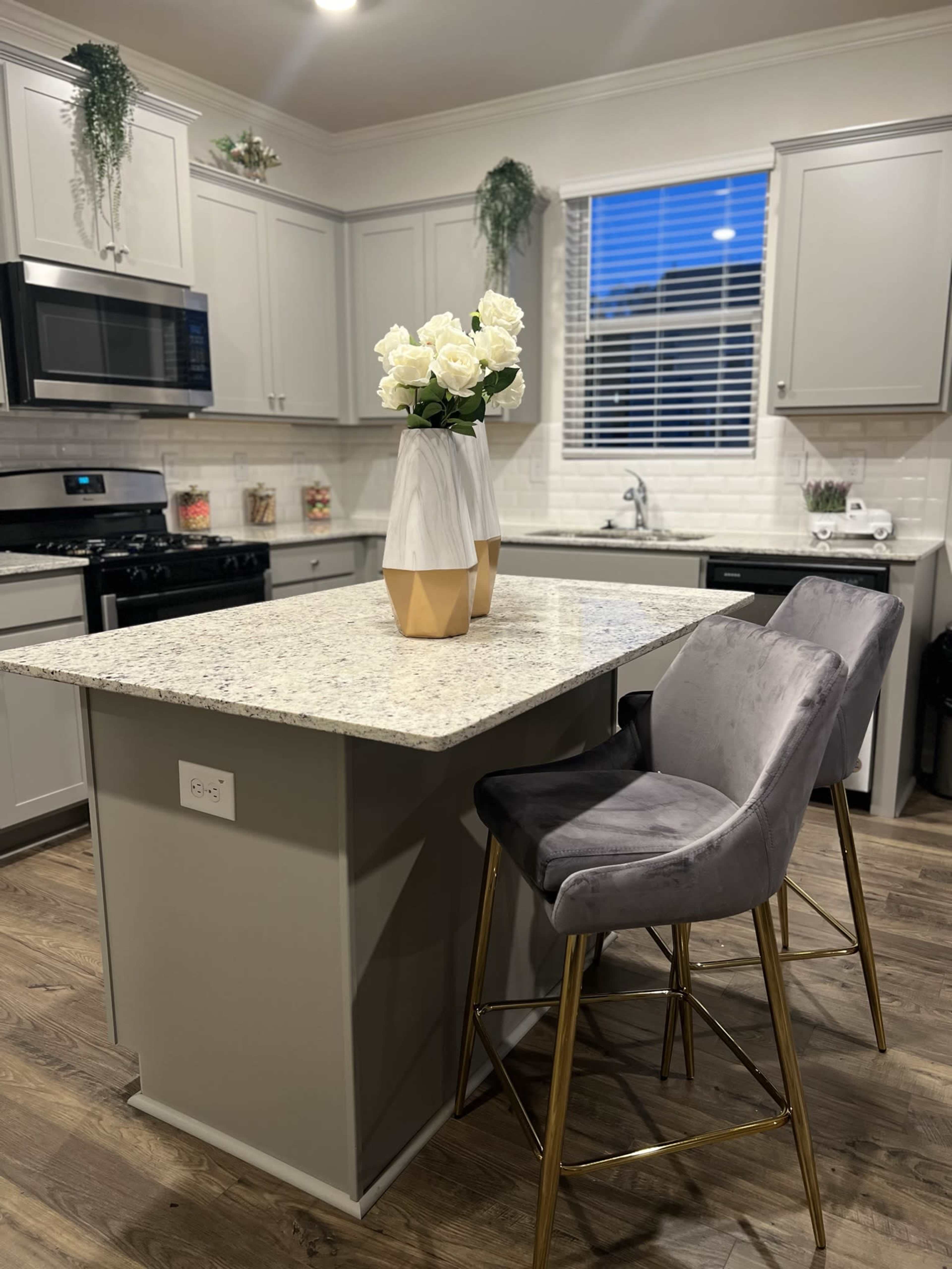 A modern kitchen features gray cabinetry, a granite island with a vase of white roses, and tall stools with gold legs.