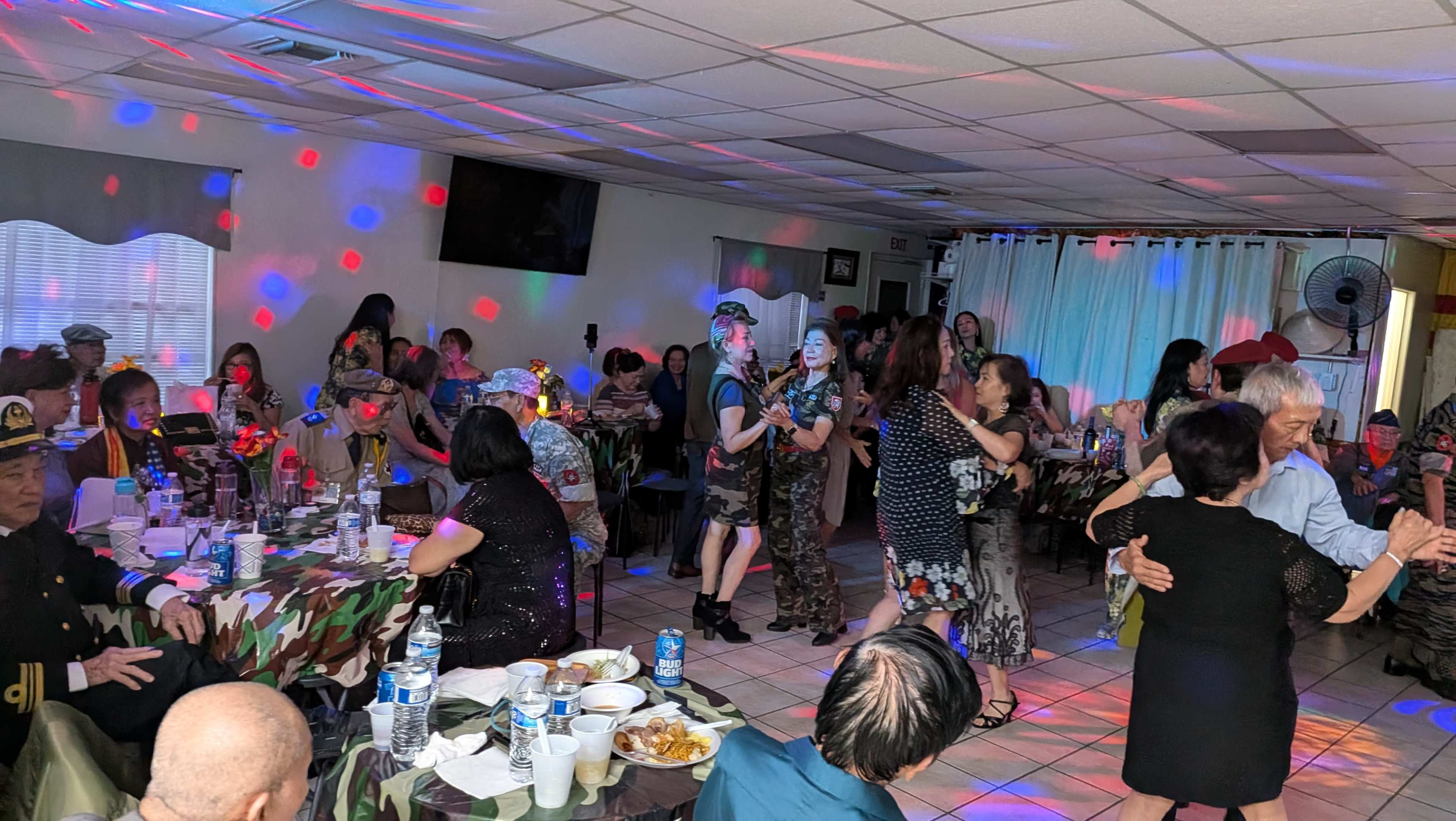 People are dancing at a gathering with colorful lights and decorated tables in a room filled with attendees.