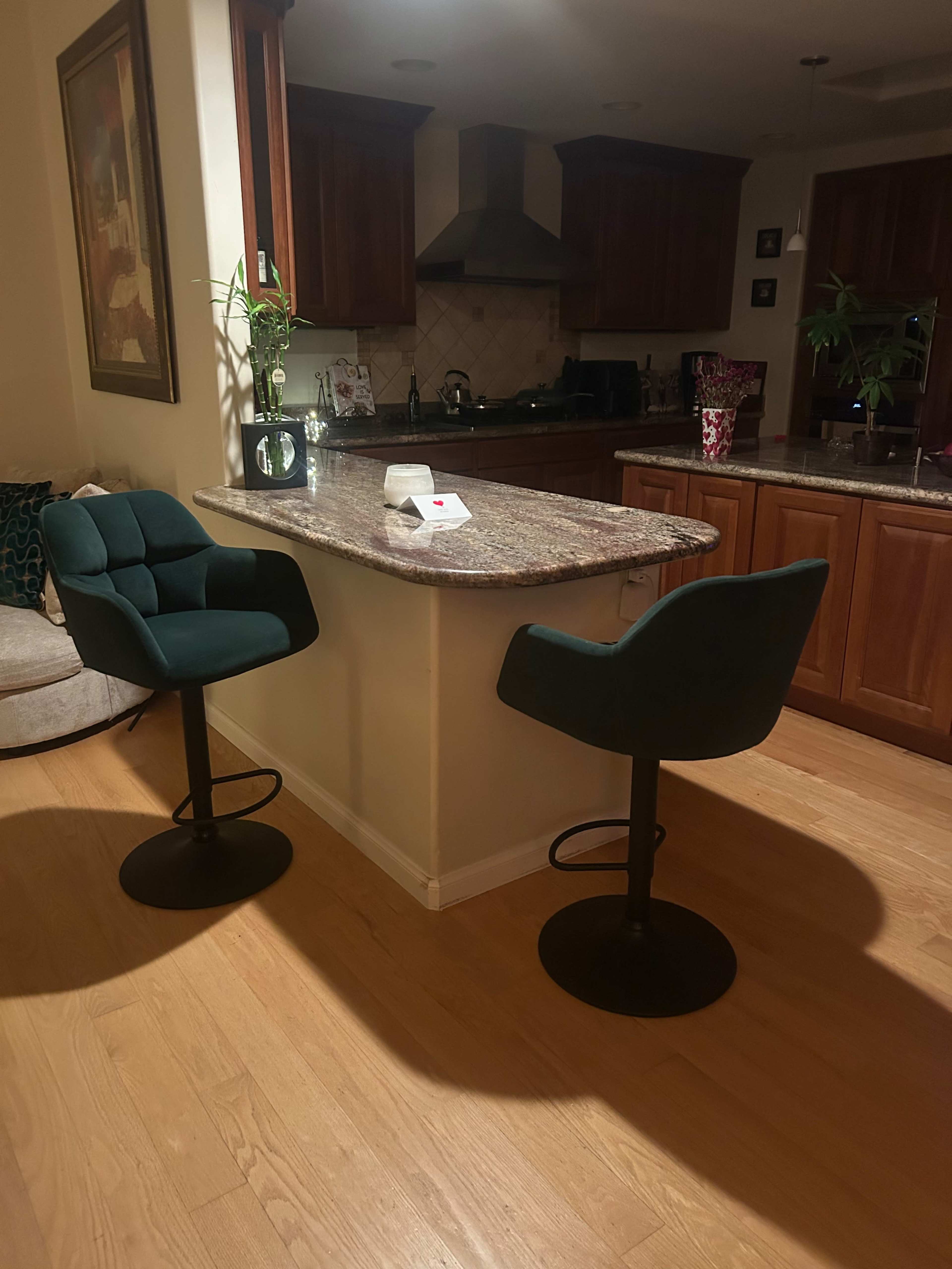 The image shows a kitchen area with two modern bar stools at a small counter, surrounded by wooden cabinetry and appliances.