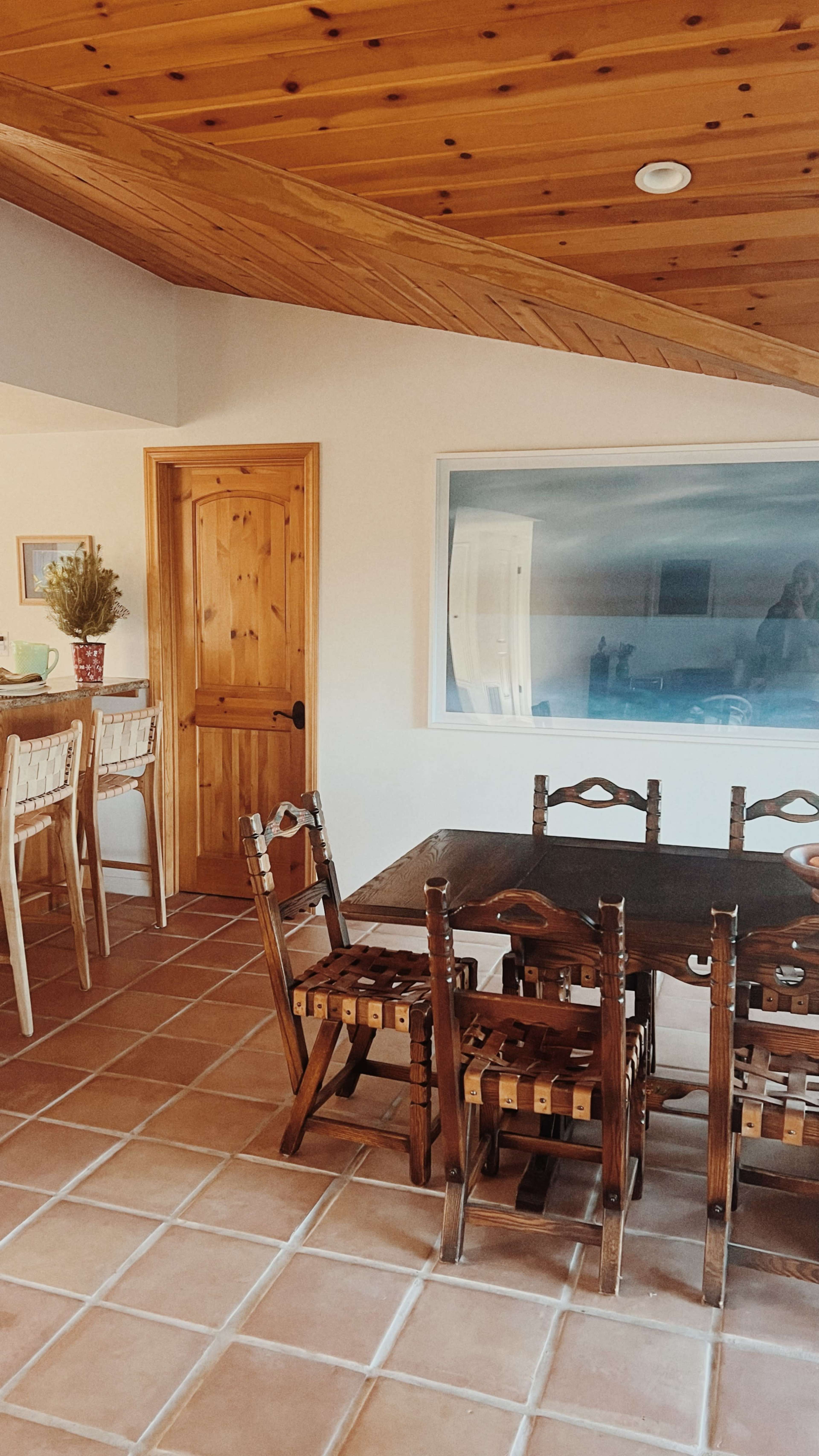 The image depicts a dining area featuring a wooden table surrounded by four chairs, with bar stools positioned at a counter and a large painting on the wall.