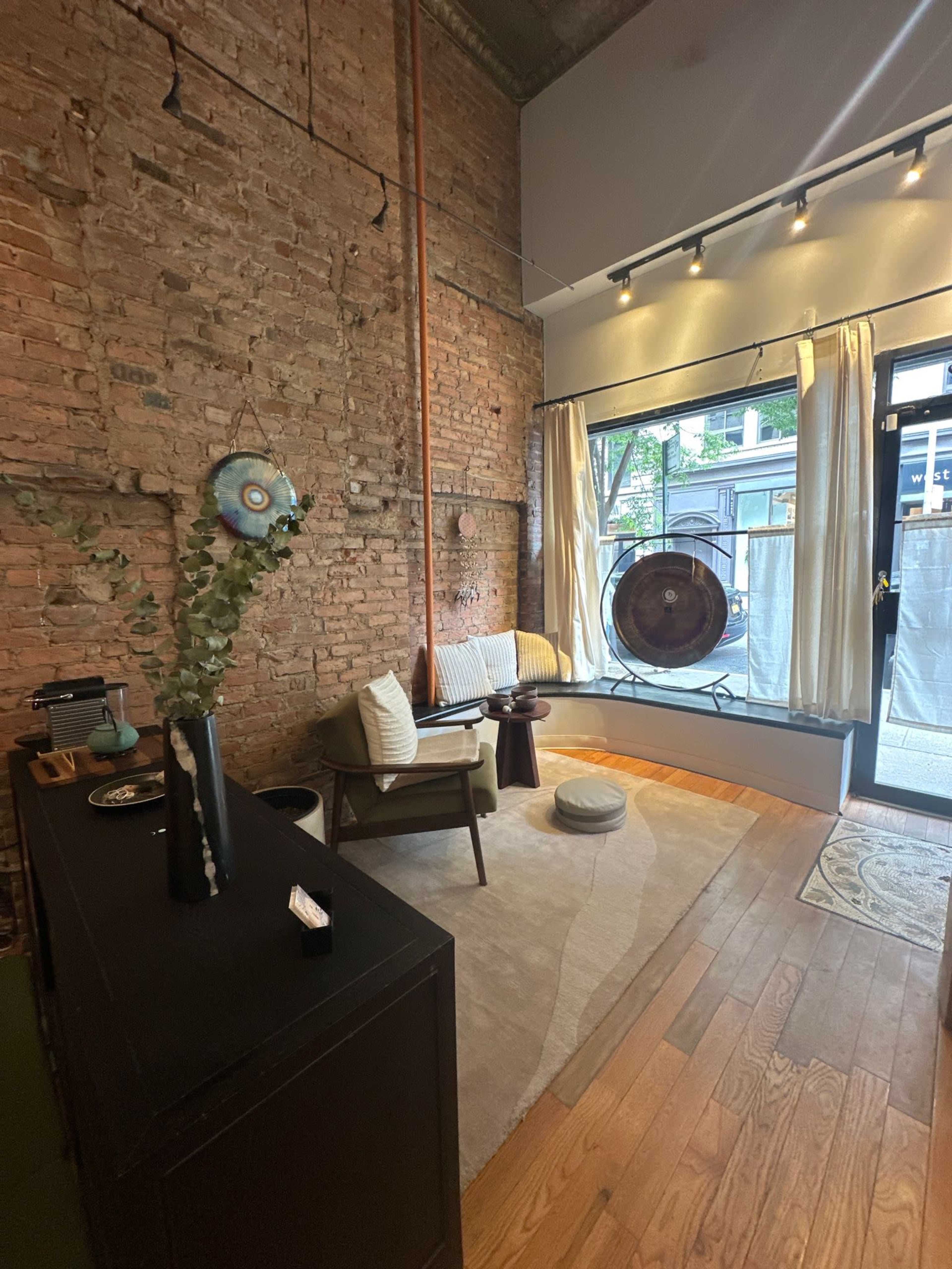 The image shows a cozy room with exposed brick walls, wooden floors, and a seating area beside a large window featuring a gong.