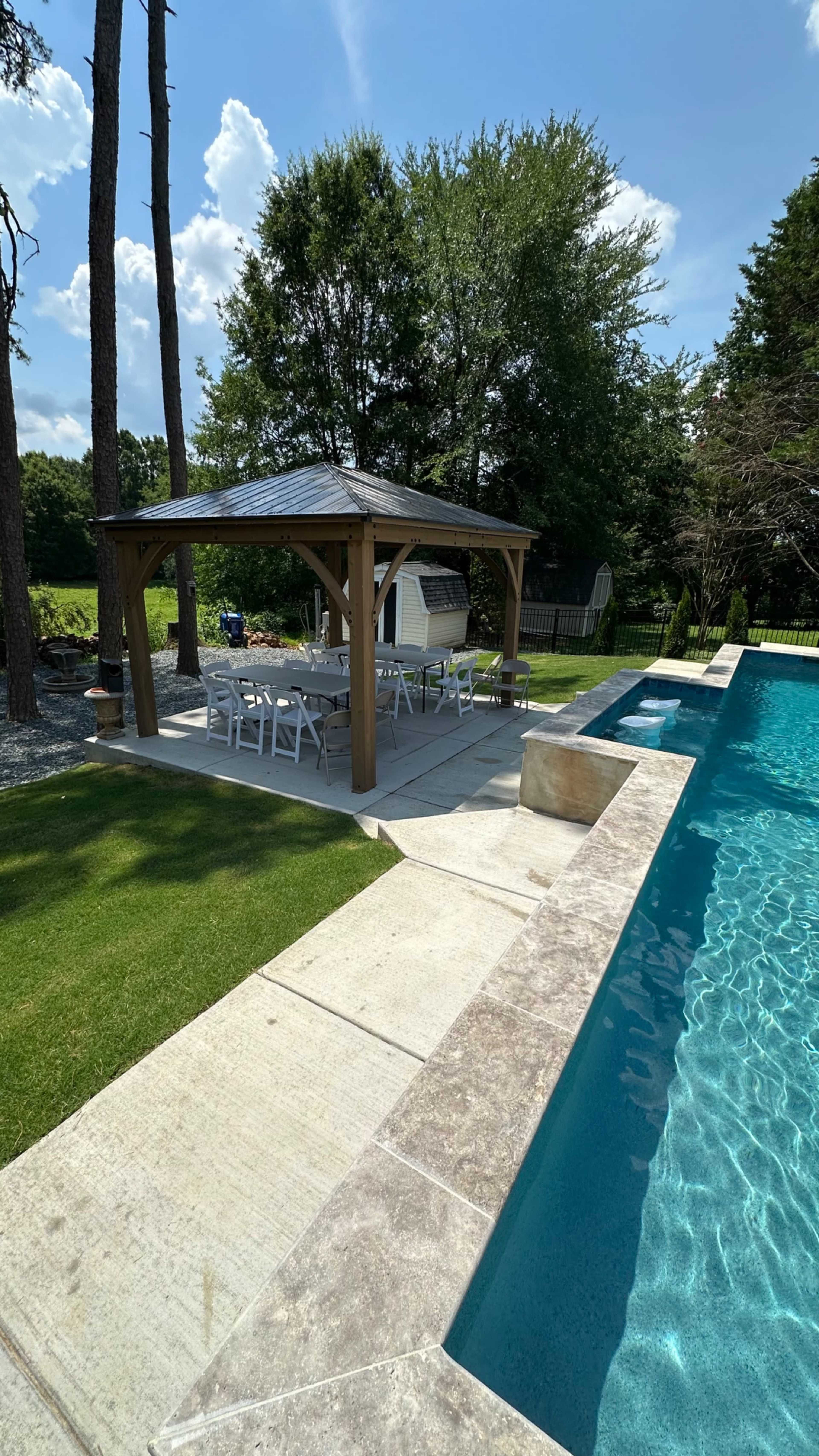 A wooden gazebo with a dining area is positioned beside a swimming pool, surrounded by grass and landscaping.