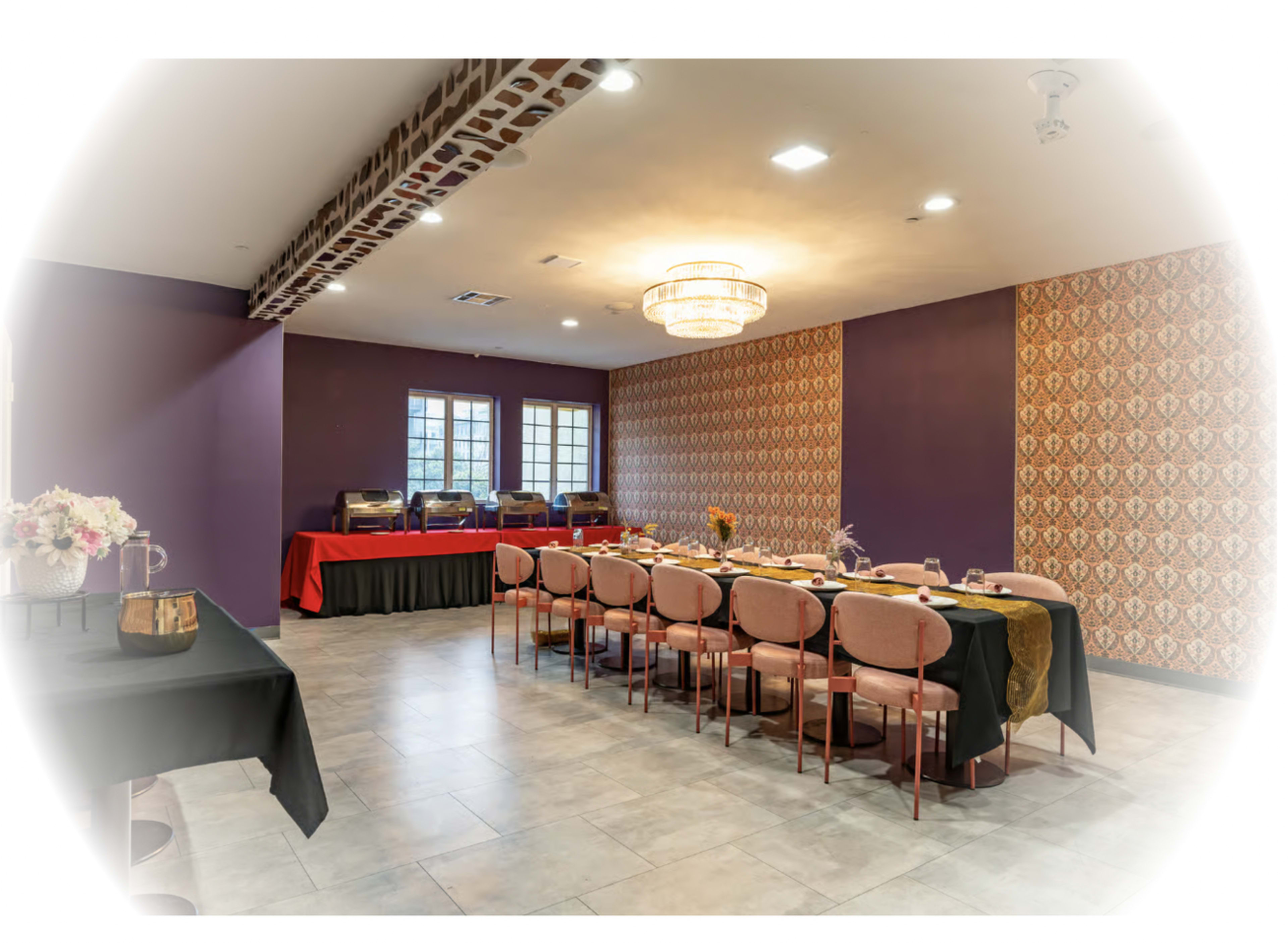 The image shows a banquet room with a long dining table set for a meal against a backdrop of decorative wallpaper and a chandelier.