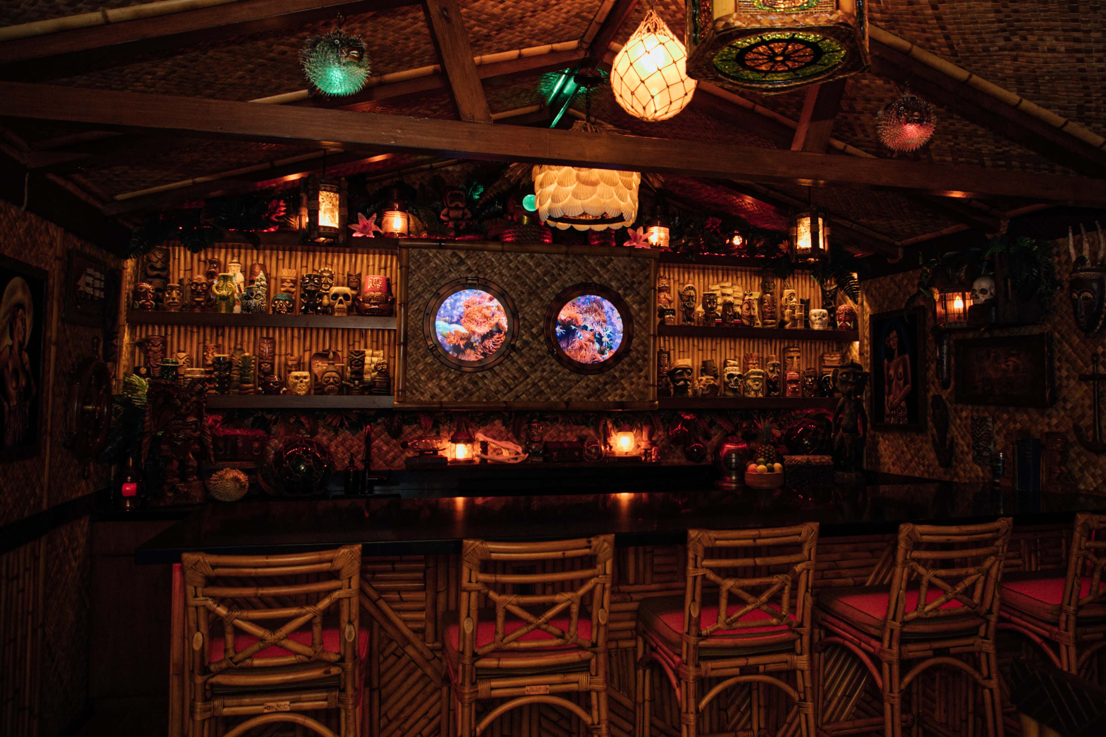 The interior of a tiki bar features bamboo decor, circular framed mirrors, and shelves lined with various decorative masks, illuminated by colorful lighting.