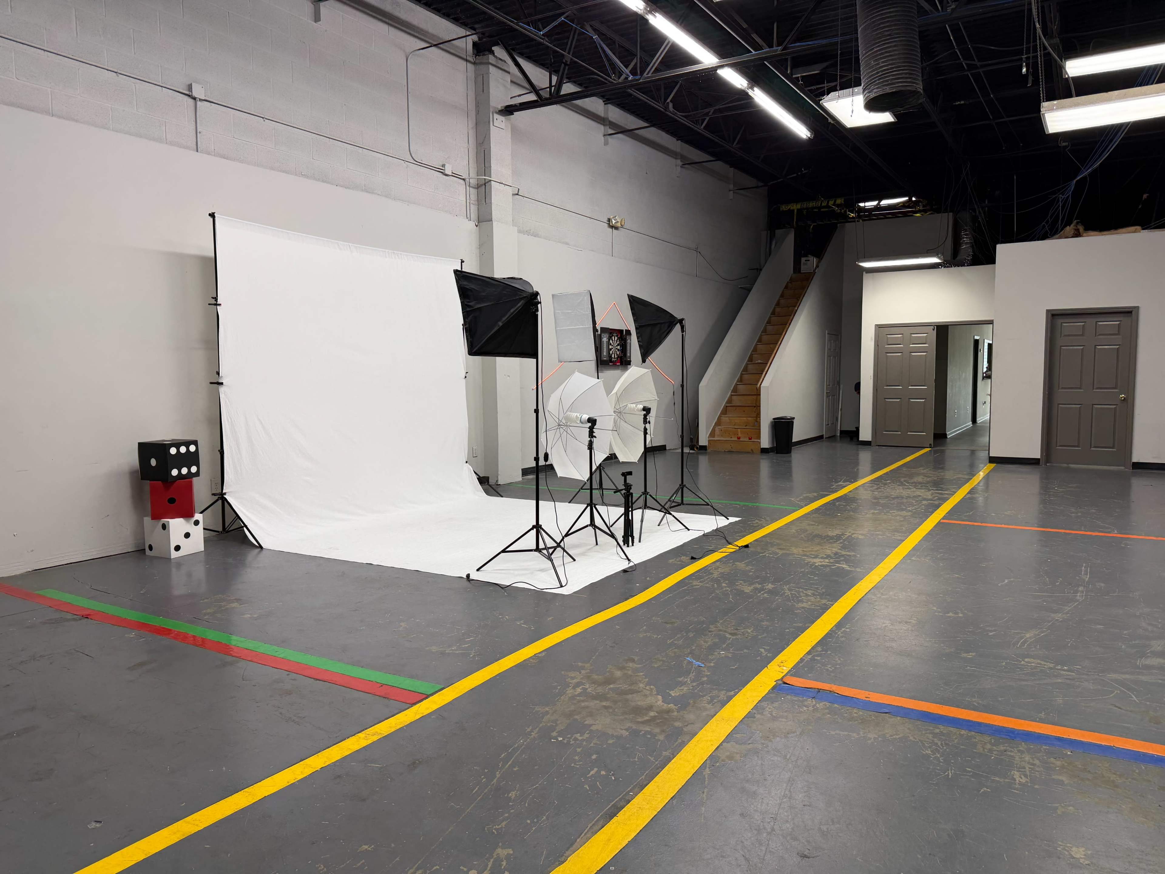 The image shows a spacious, well-lit studio setup with a white backdrop, lighting equipment, and a staircase in the background.