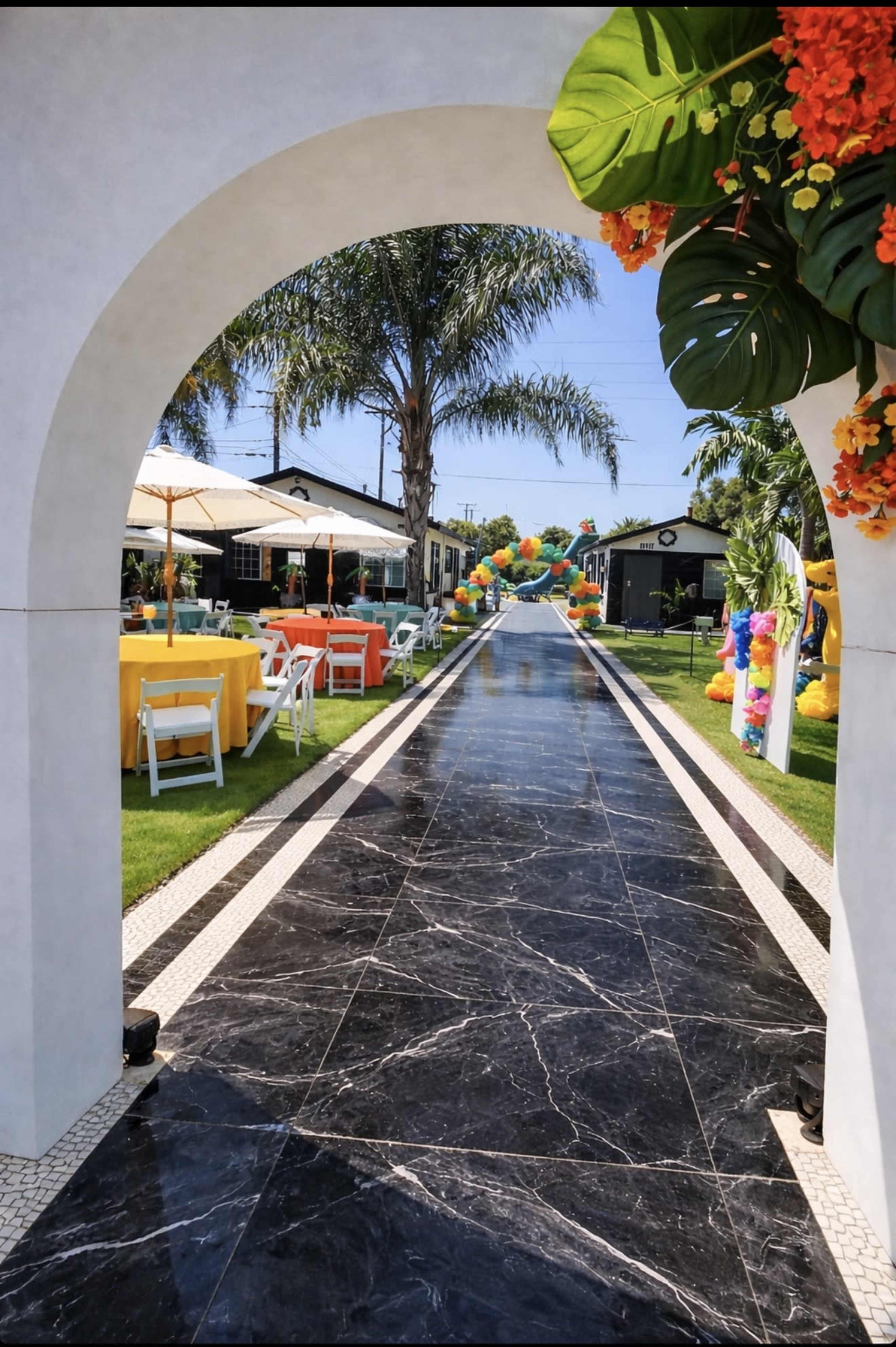 The image shows a pathway lined with black marble tiles, leading through an archway into an outdoor area adorned with colorful decorations and tables set for an event.