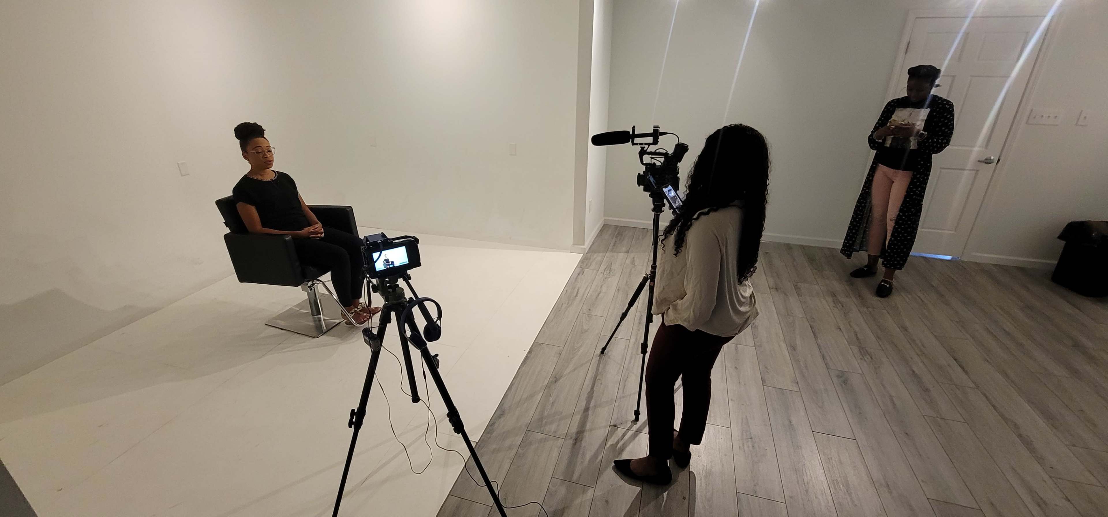 A person sits in a chair during an interview while another person operates a camera, and a third person stands in the background holding a clipboard.