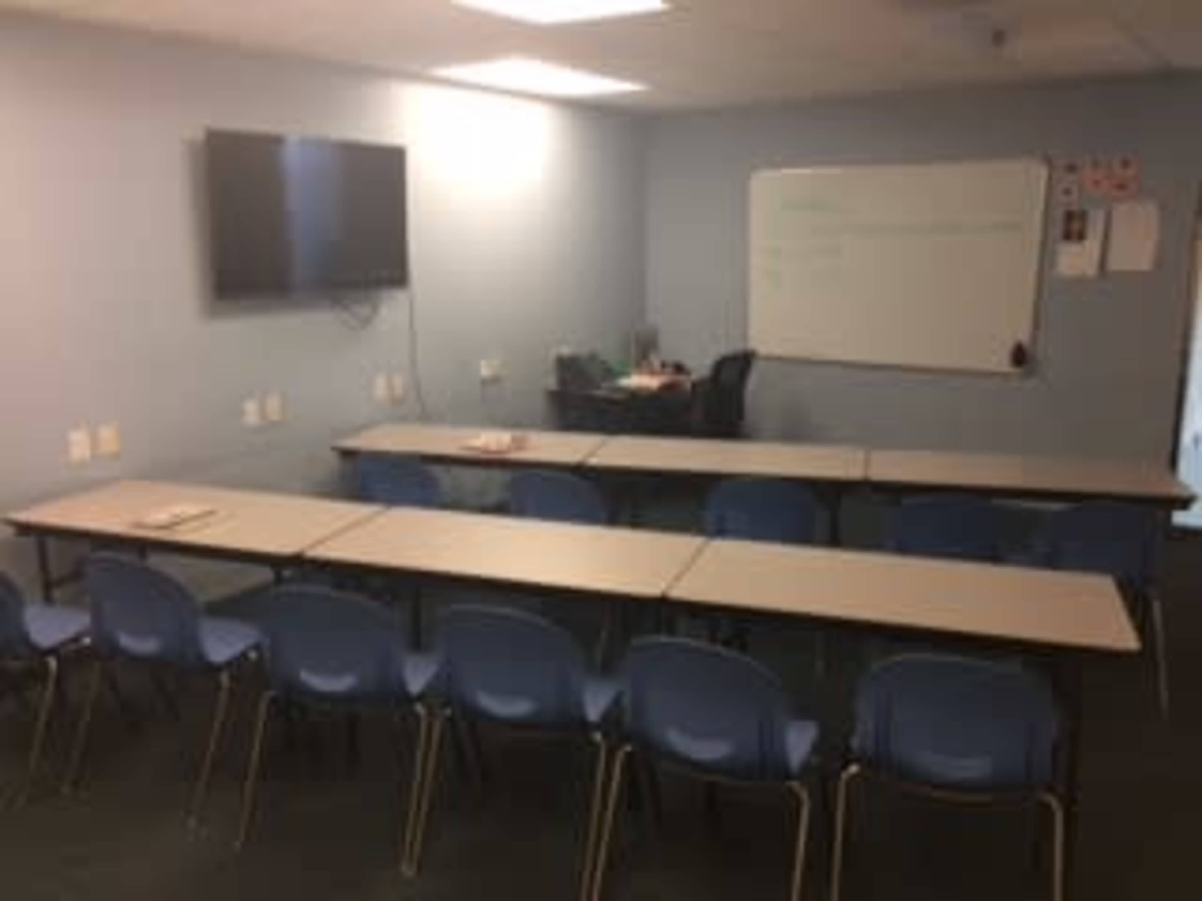 The image shows a classroom setup with multiple tables arranged in a U-shape and blue chairs, along with a whiteboard and a television on one wall.
