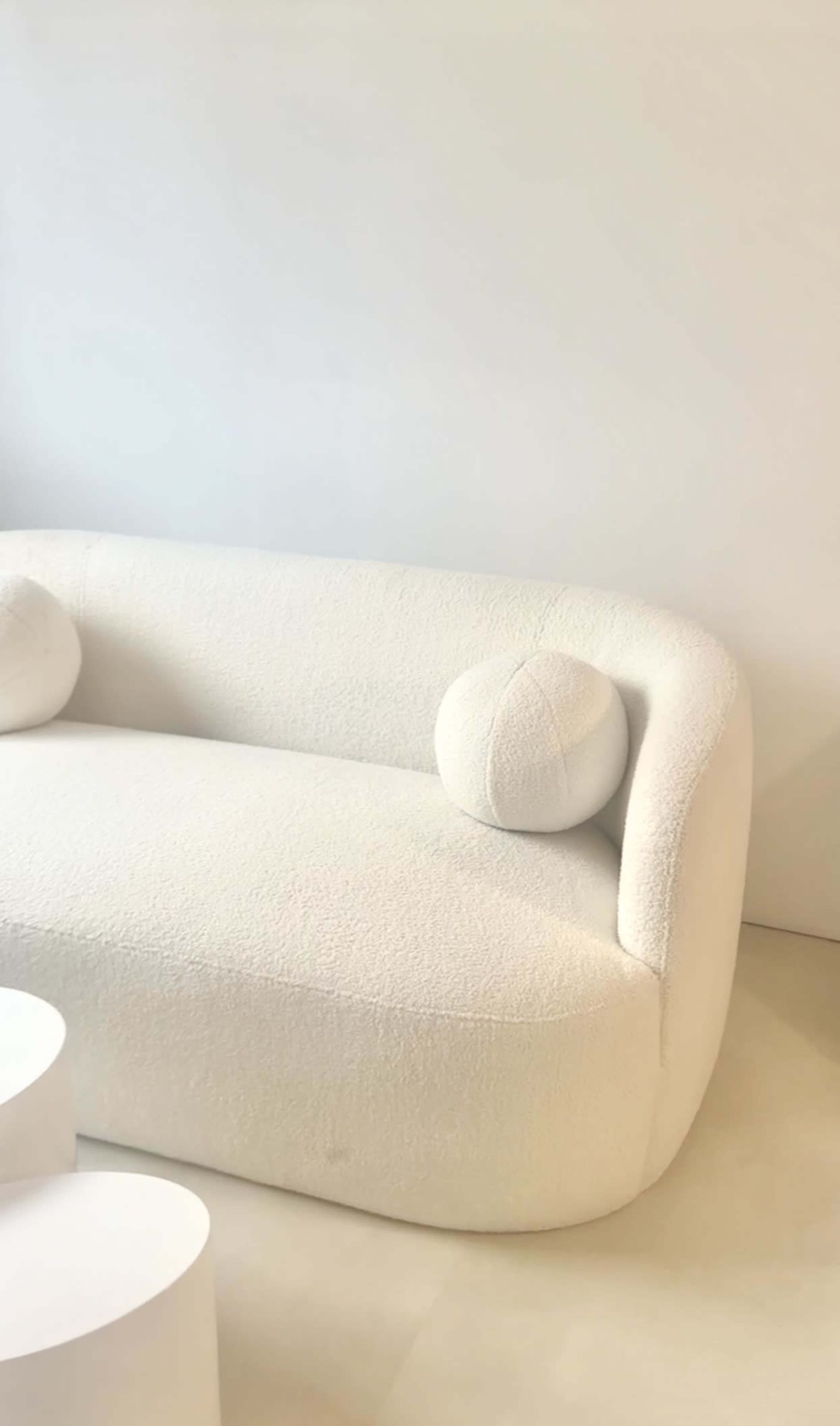 The image shows a white, upholstered sofa with rounded edges and a circular cushion on either end, set against a plain white wall.