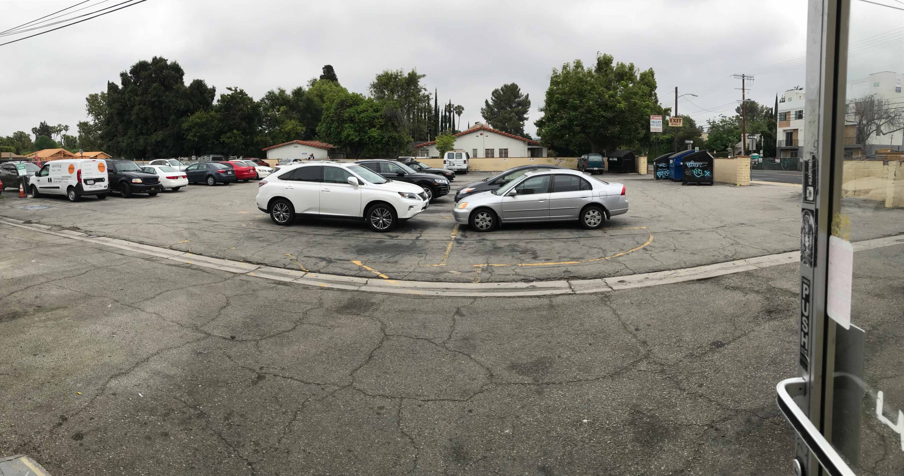 The image shows a parking lot filled with vehicles, including a white SUV and a silver sedan in the foreground, surrounded by trees and buildings.
