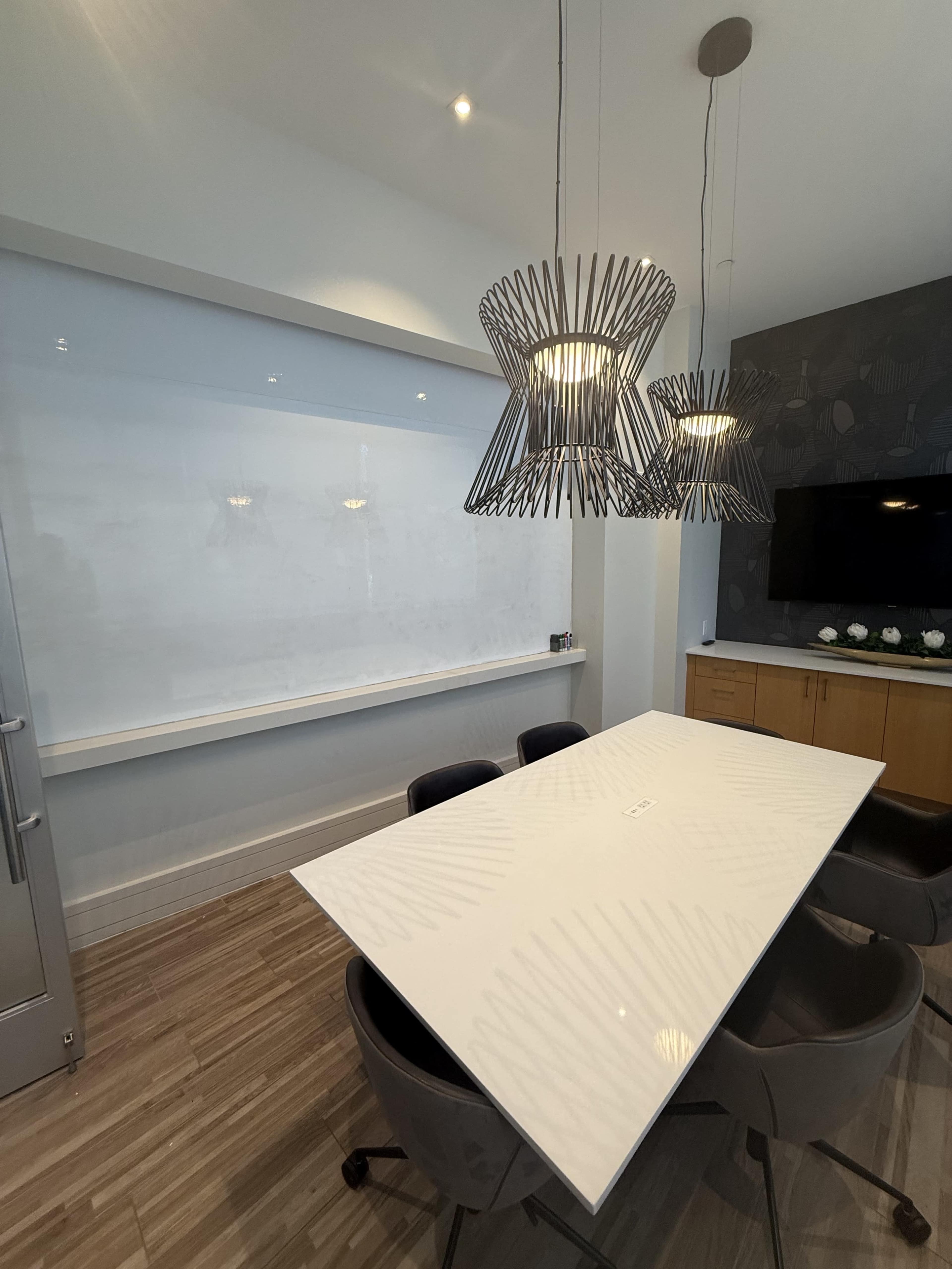 A modern conference room features a white table, grey chairs, decorative pendant lights, and a large whiteboard on the wall.