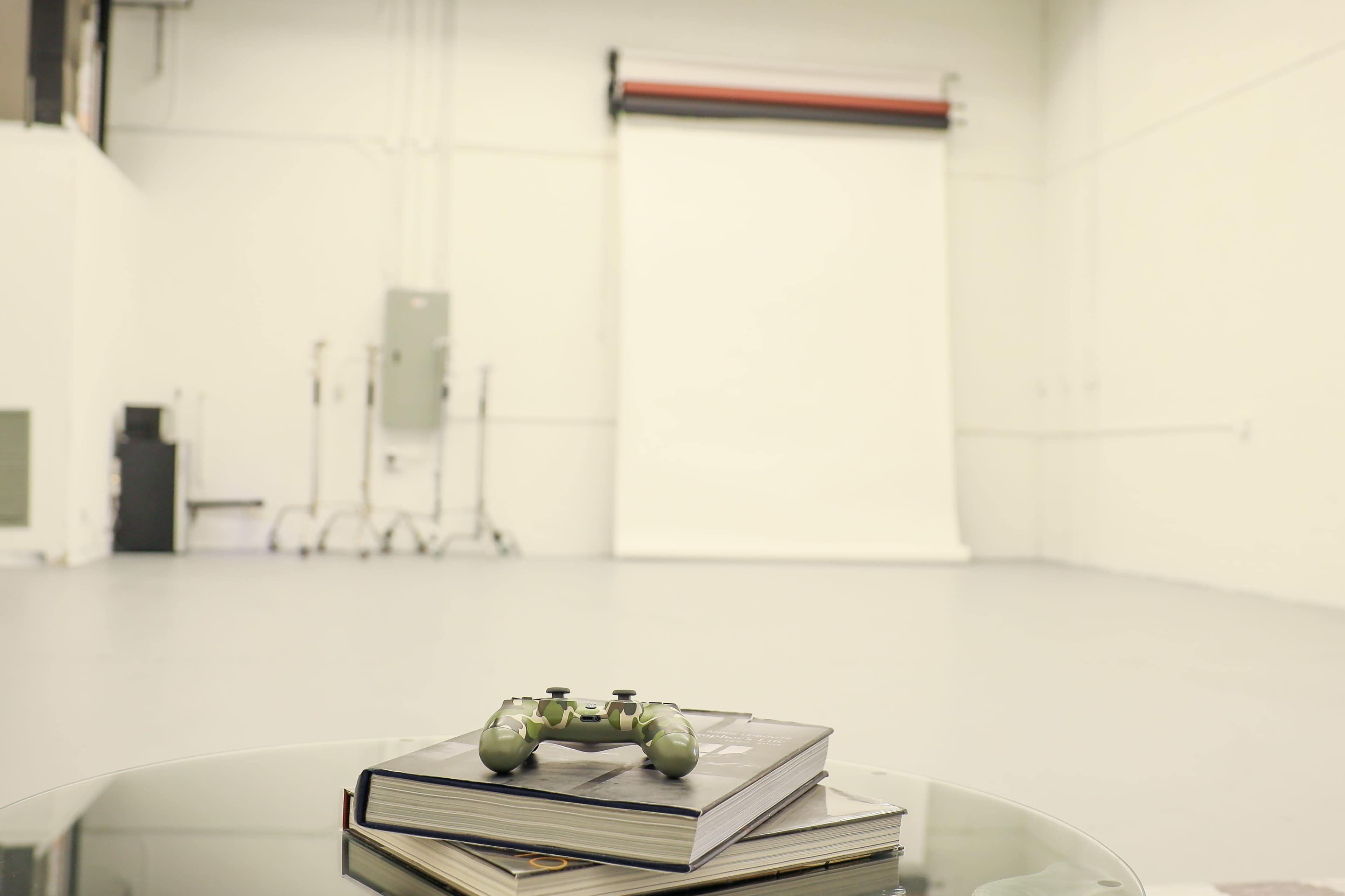 A green video game controller rests on top of two stacked books in an empty studio space with a white backdrop.