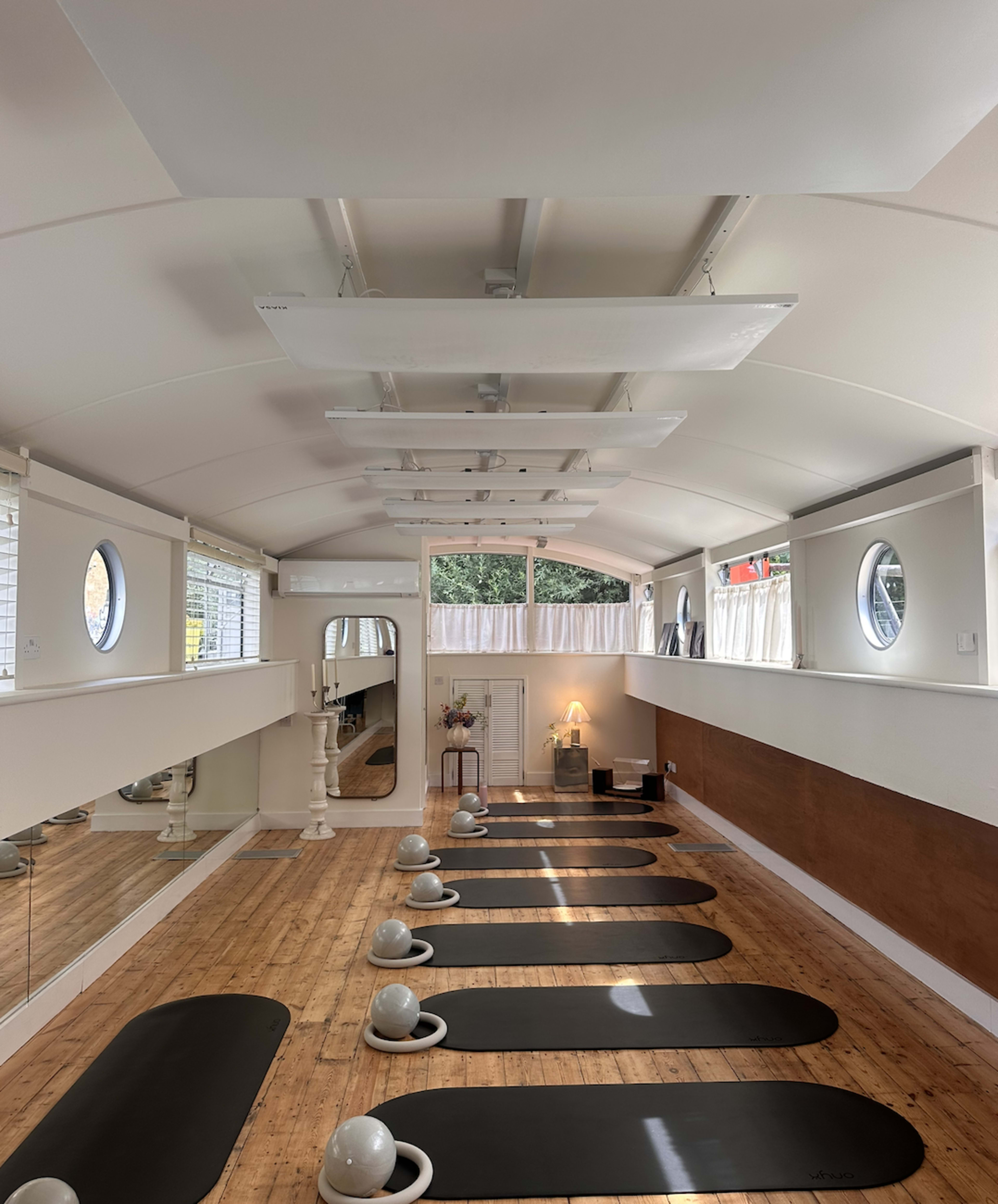 The image shows a minimalist yoga studio with several black mats arranged neatly on a wooden floor, illuminated by natural light through large windows.