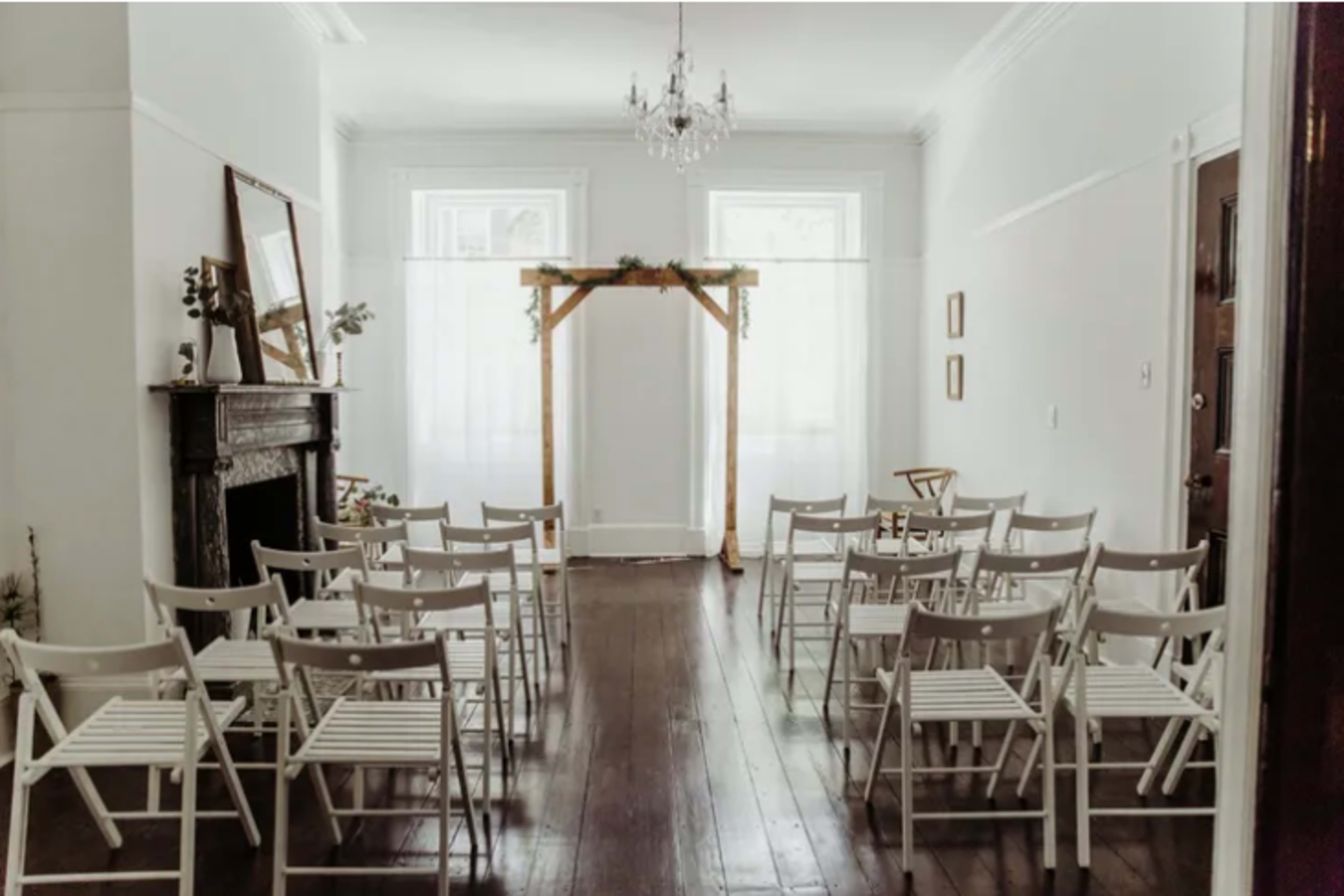 The room features a simple setup with white folding chairs arranged in rows facing a wooden archway, with a chandelier hanging above and light streaming through the windows.