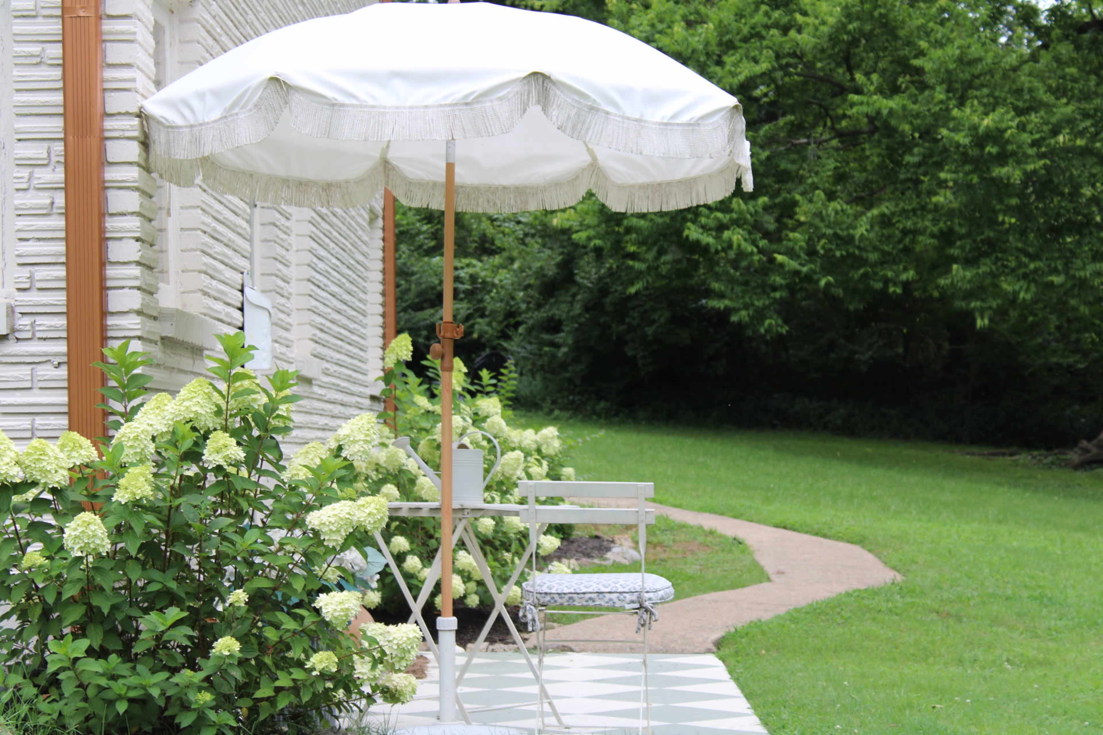 A white umbrella stands over a small table and chair set surrounded by green foliage and a path leading through the grass.