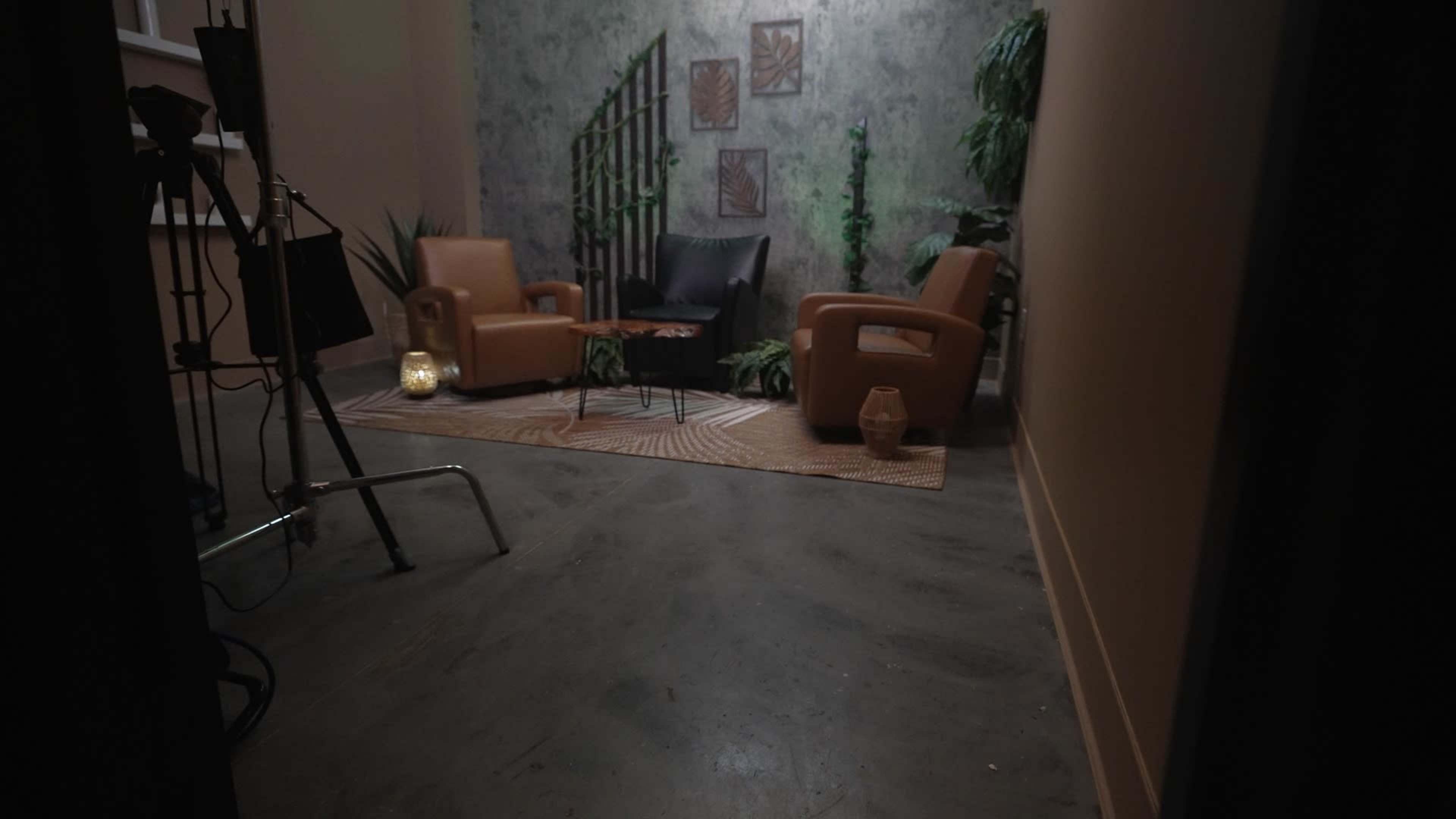 The image shows a small indoor seating area with two brown chairs, a table, and decorative plants against a textured wall.