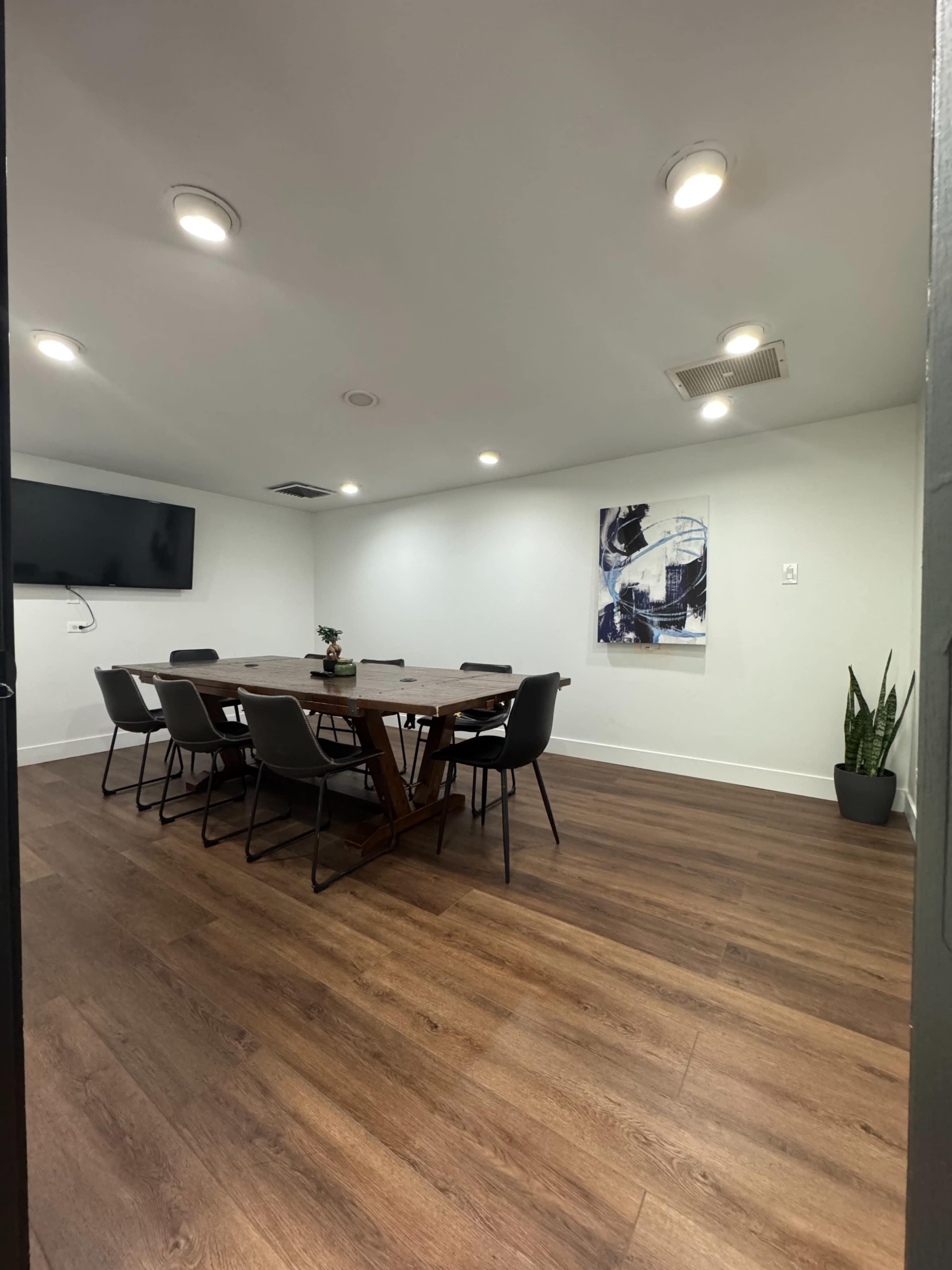 A modern conference room features a large wooden table surrounded by eight chairs, with a television mounted on one wall and a plant in the corner.