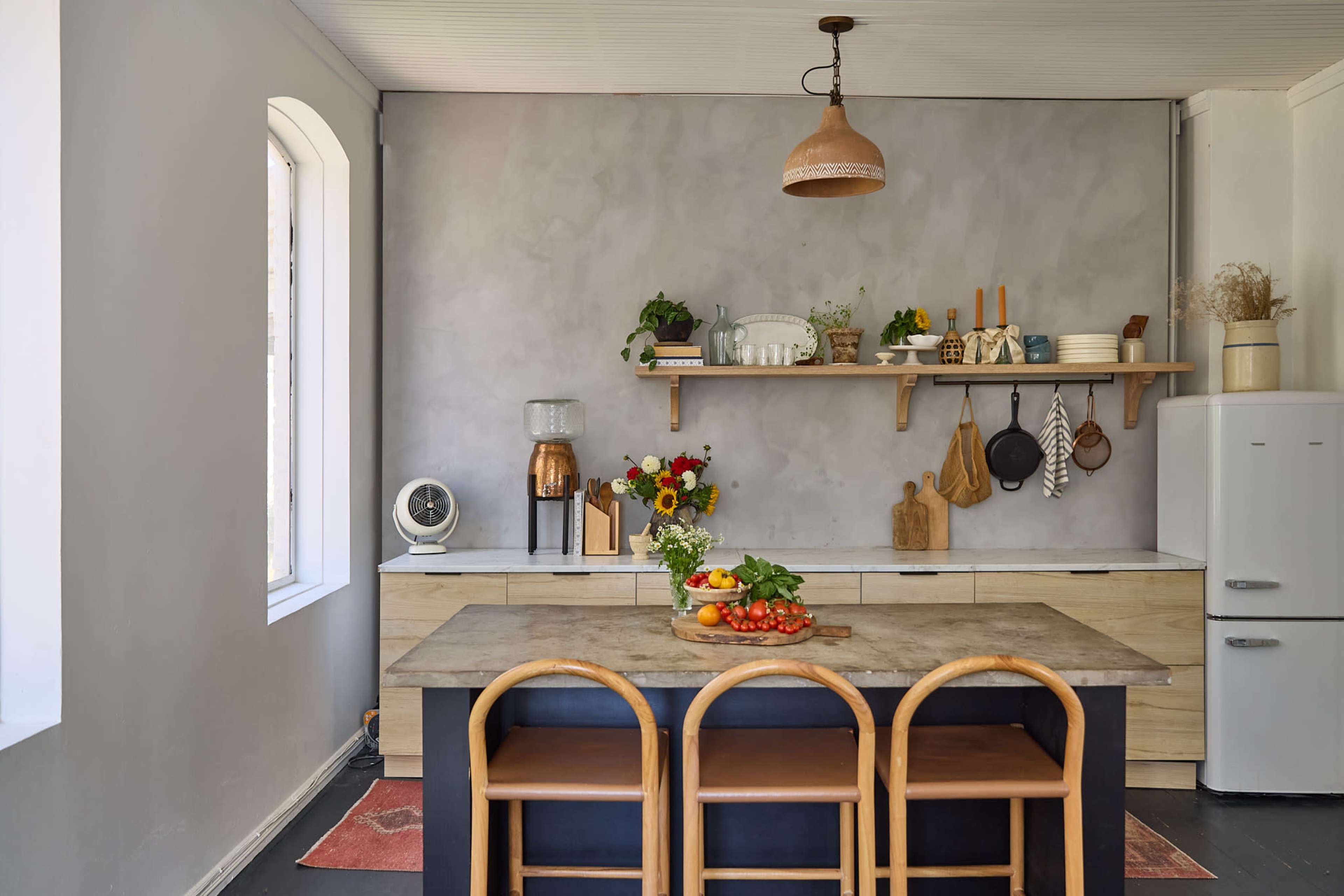 A modern kitchen features a wooden table with three rattan chairs, a wall shelf with plants and kitchenware, and a white refrigerator in the corner.