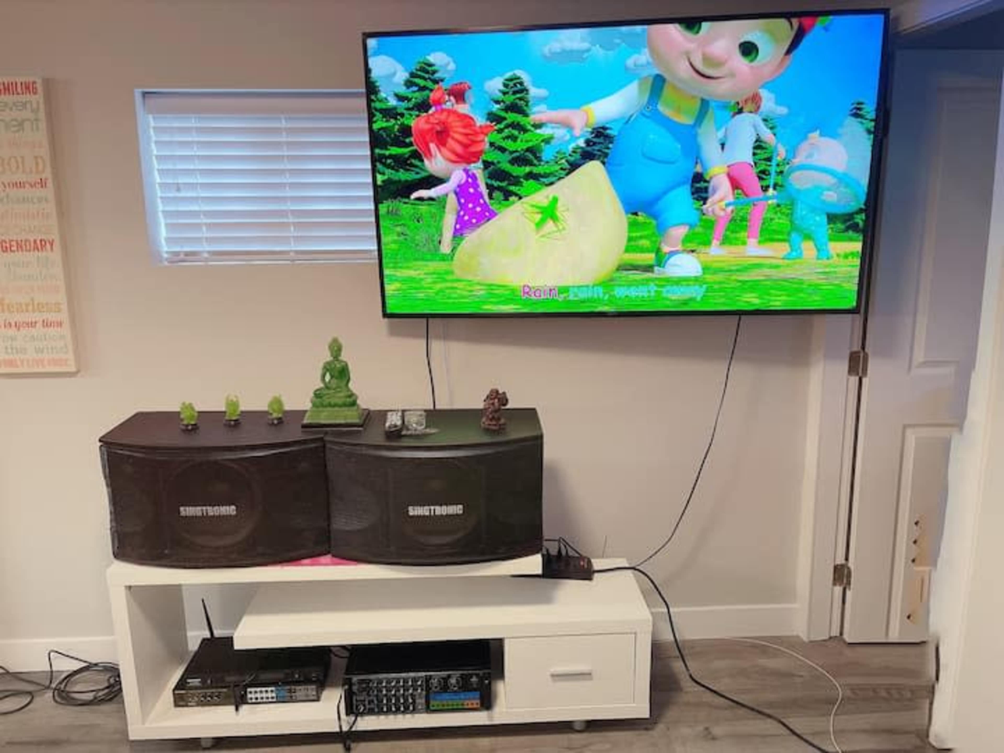 A wall-mounted television displays an animated children's show, while a white entertainment unit beneath holds speakers and various decorative items.