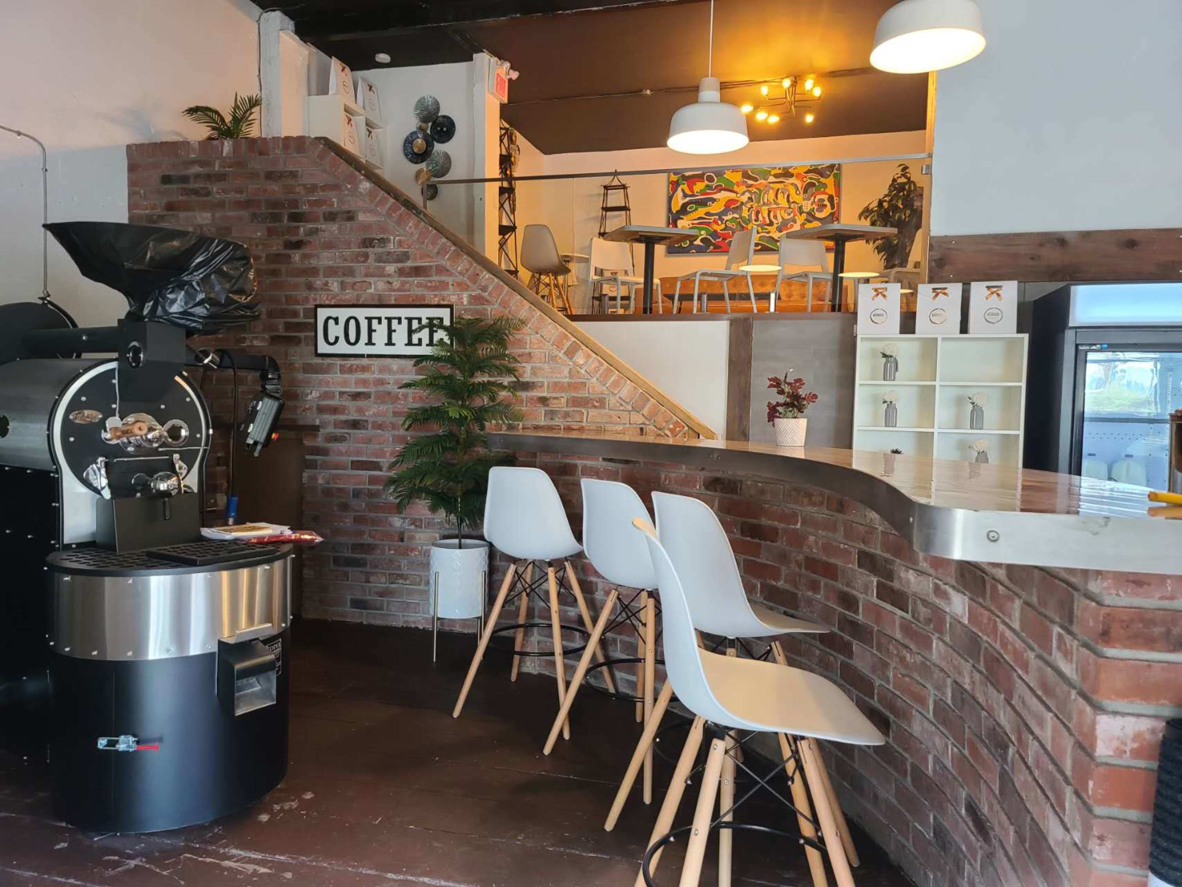 A coffee shop with a bar area featuring three white stools, a coffee roasting machine, and a staircase leading to an upper seating area.