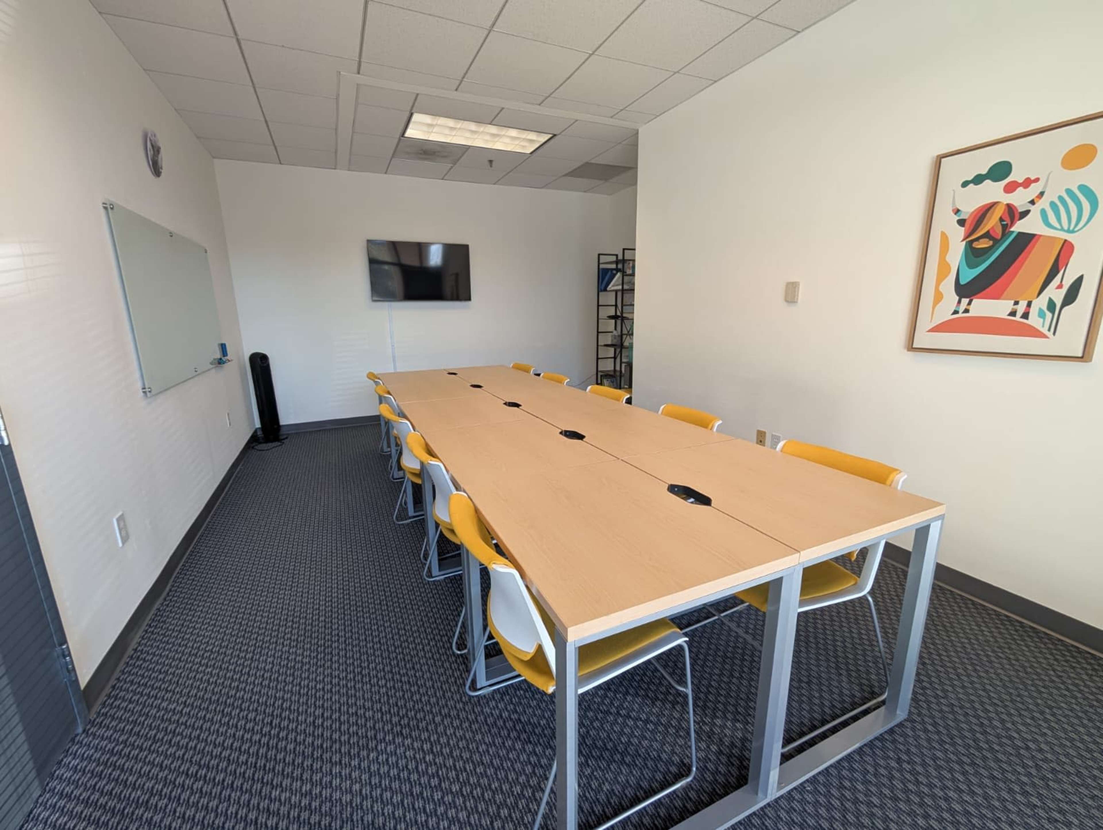 A meeting room features a long wooden table surrounded by yellow chairs, a wall-mounted TV, and a whiteboard.