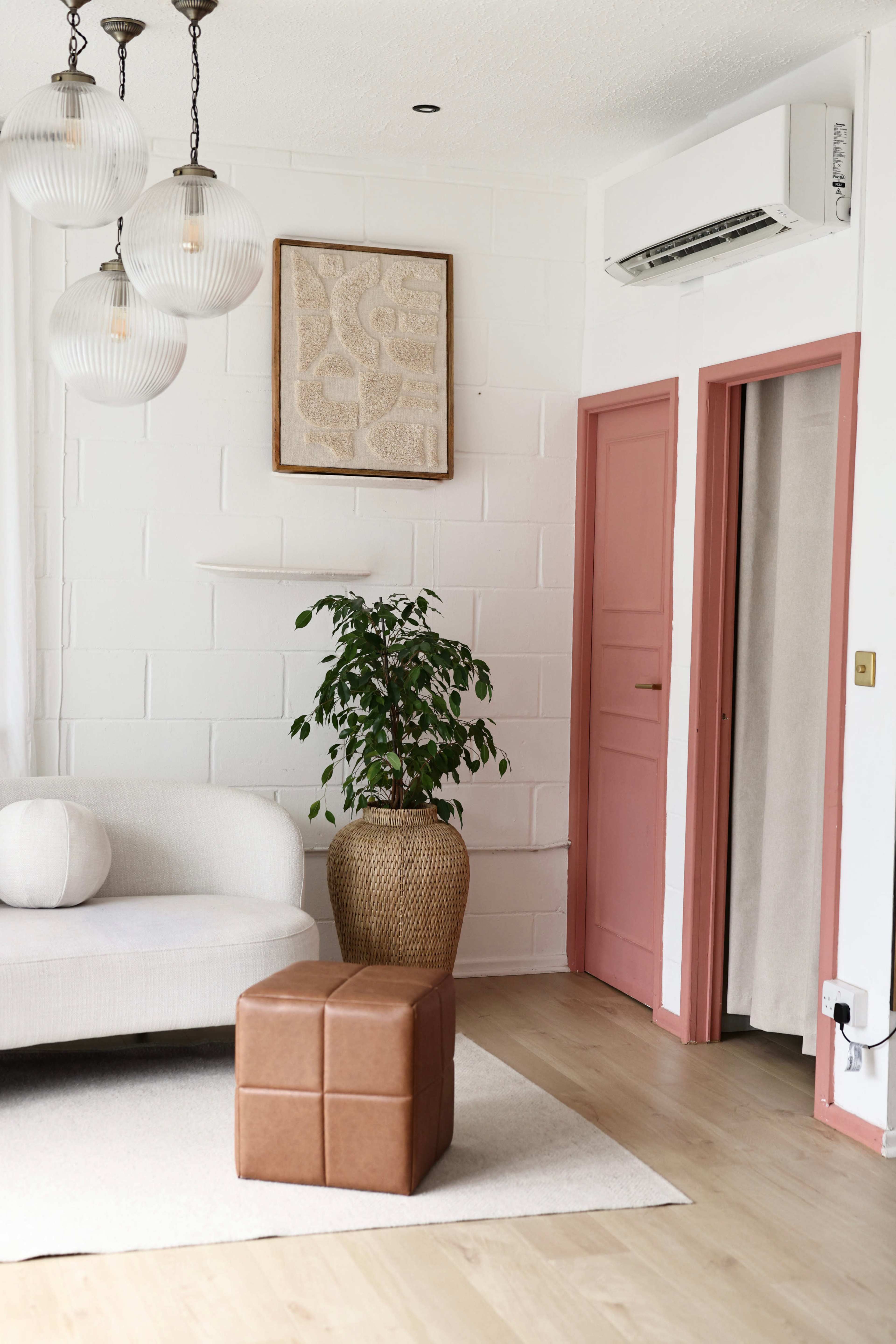 The image shows a modern living room with a light-colored sofa, a brown leather ottoman, a potted plant, and a decorative wall hanging, complemented by pastel-colored doors.