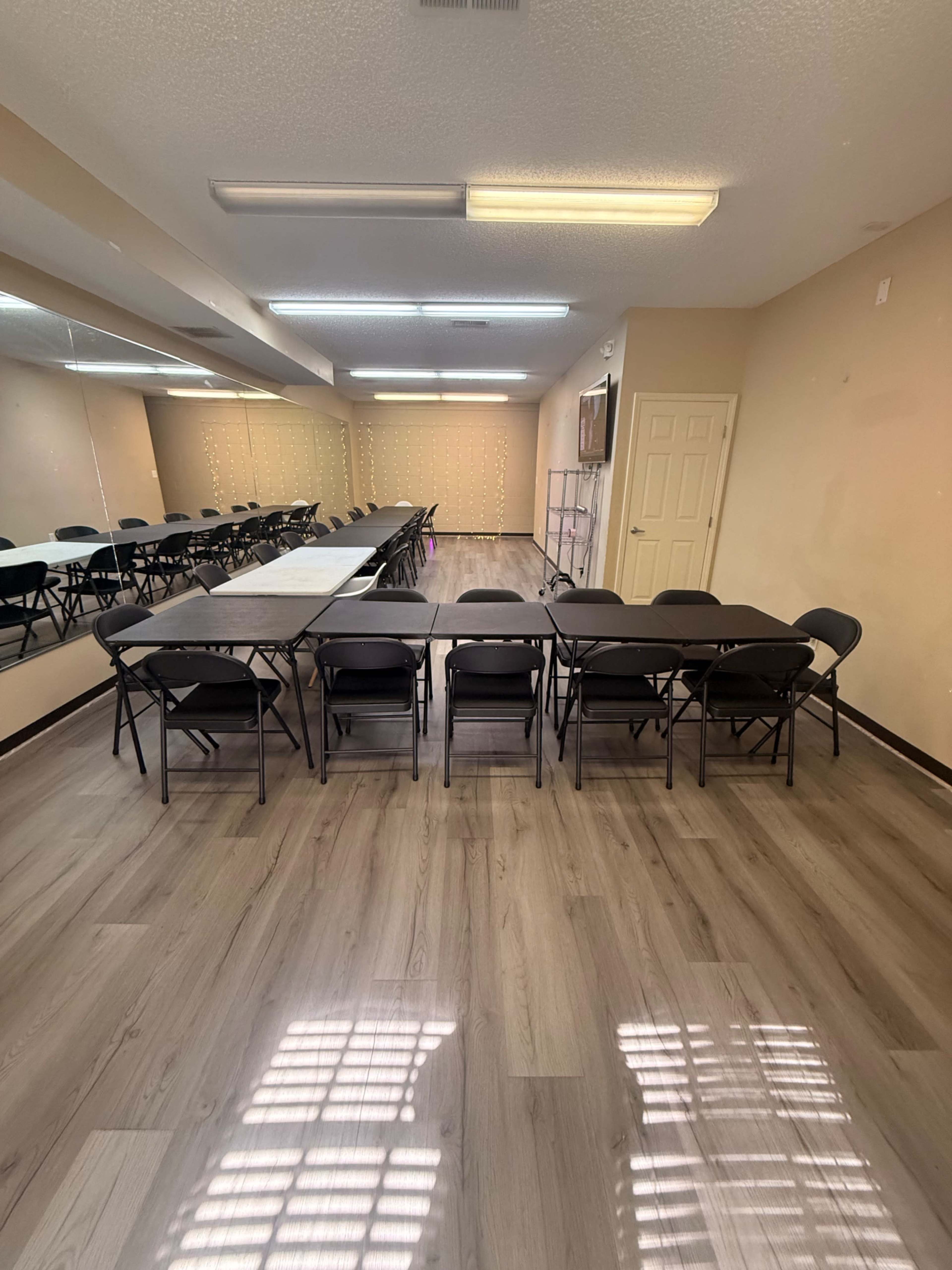 The image shows a spacious room with several rows of black folding chairs arranged around long tables, and a large mirror on one wall.