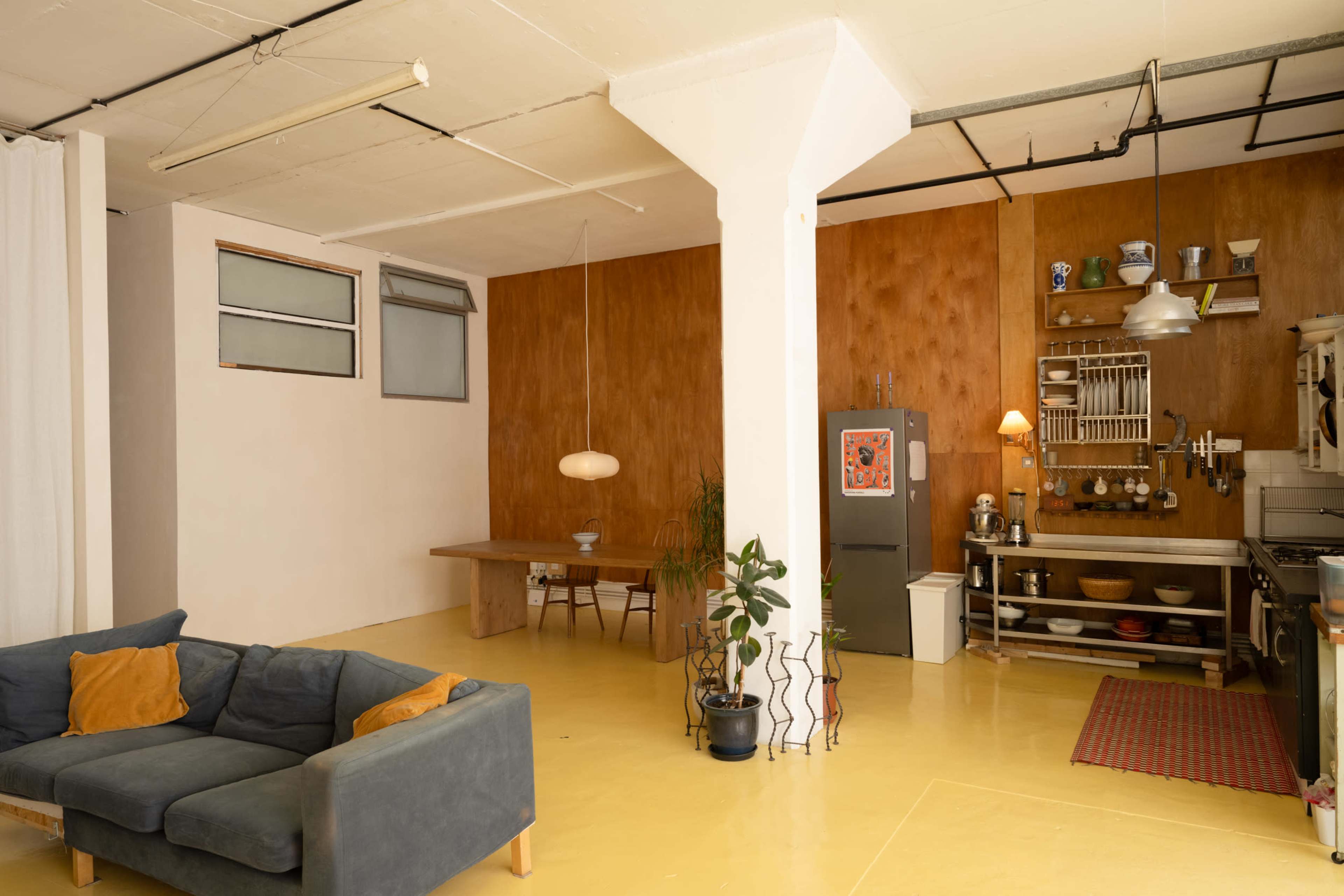 The image shows a spacious, minimalist interior featuring a living area with a couch and a kitchen area with wooden accents and simple furnishings.