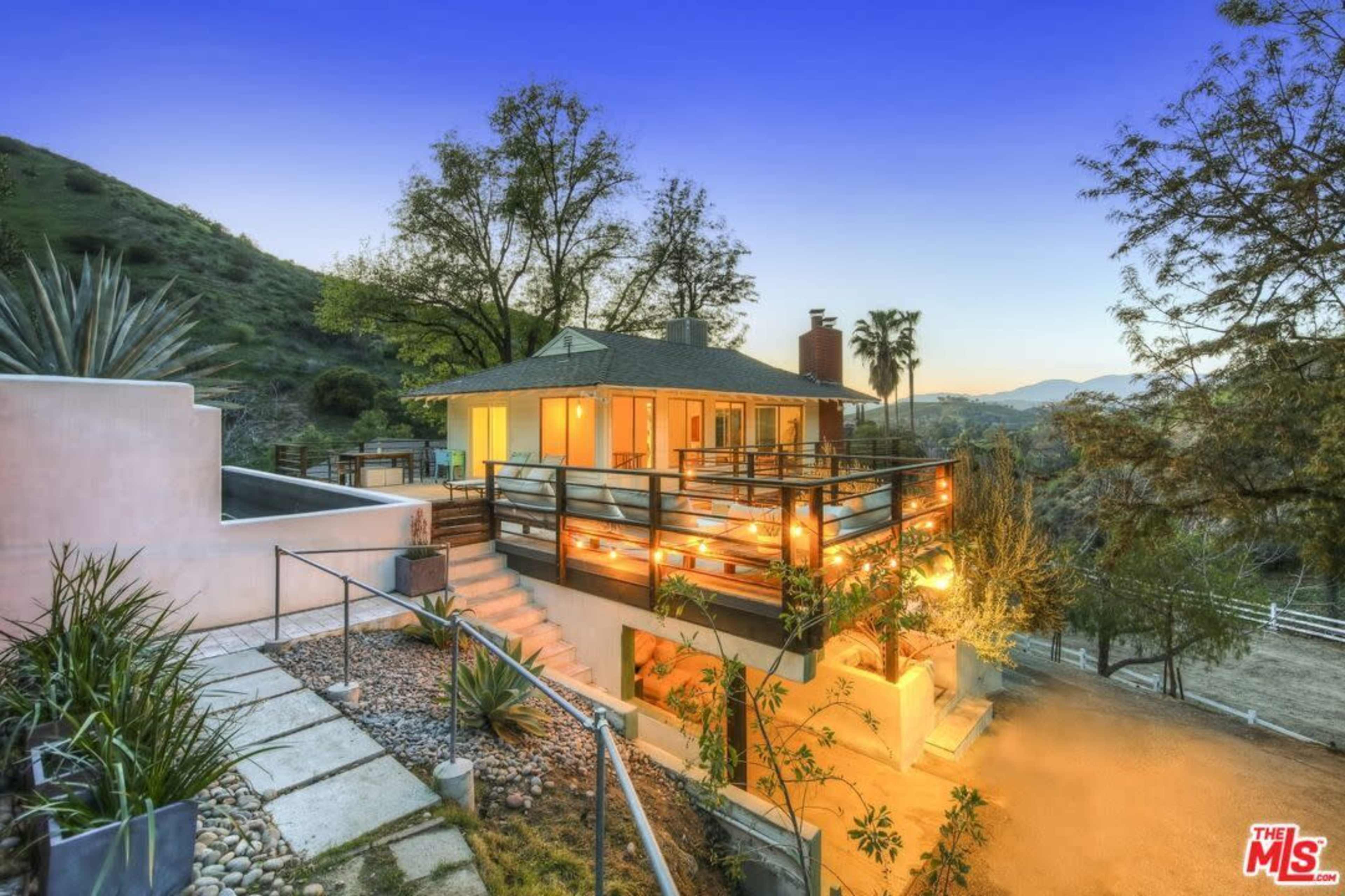 A modern house situated on a hillside, surrounded by greenery and visible outdoor lighting, with a pathway leading to the entrance.
