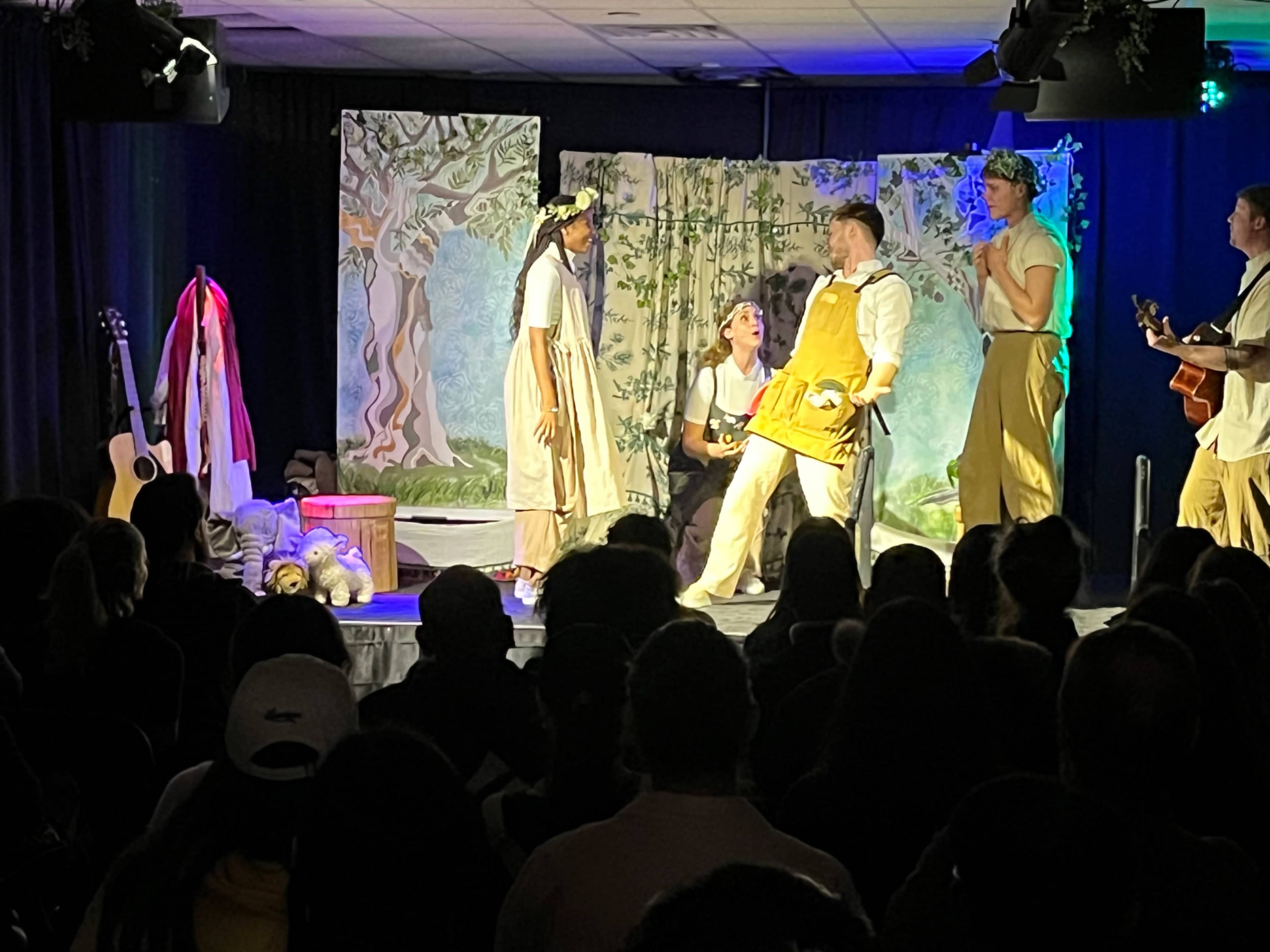 A group of five performers is presenting a theatrical play on stage, surrounded by a set with painted backdrops and props, while an audience watches attentively.