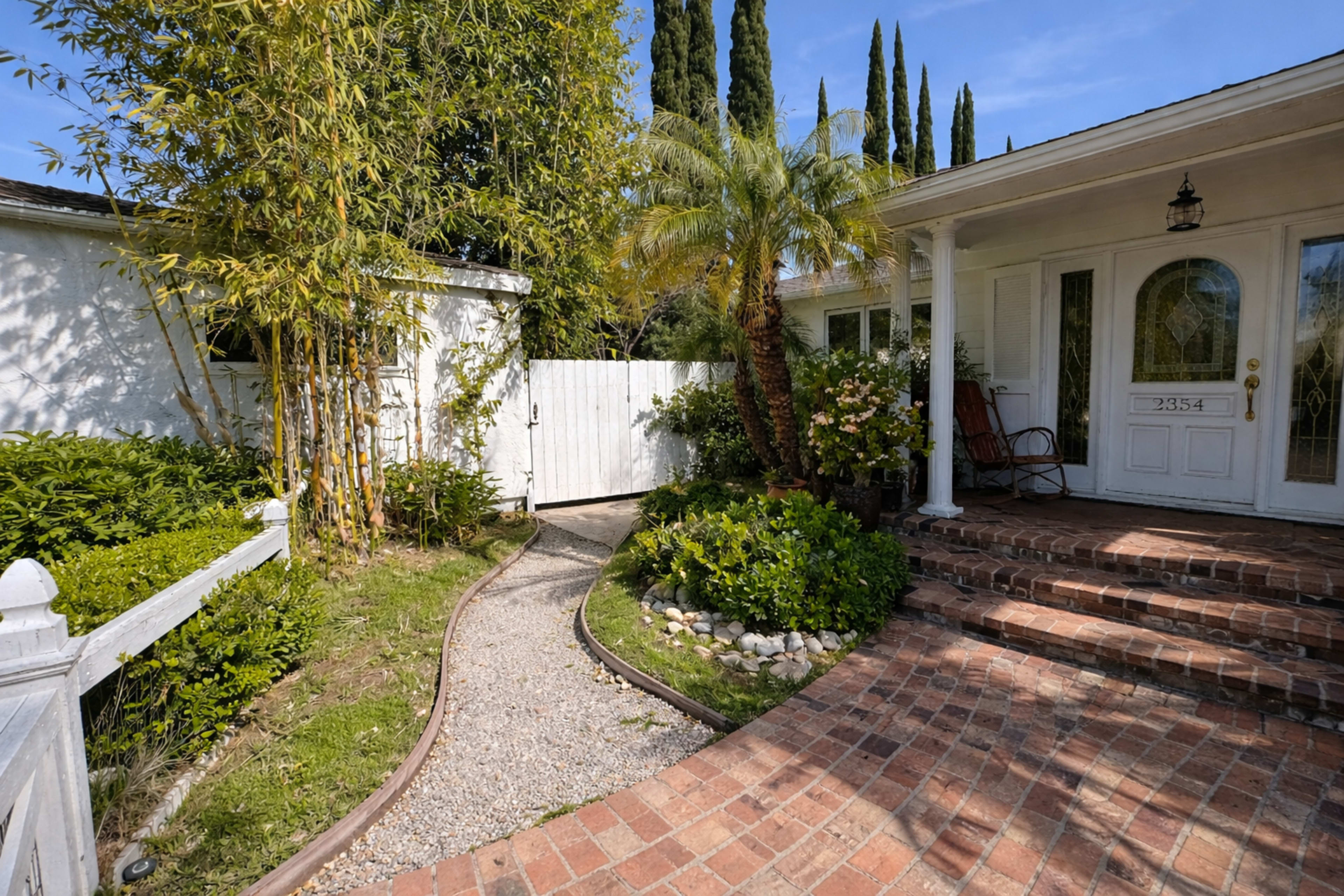 A winding gravel path leads to a white front door surrounded by greenery and potted plants, with tall trees in the background.