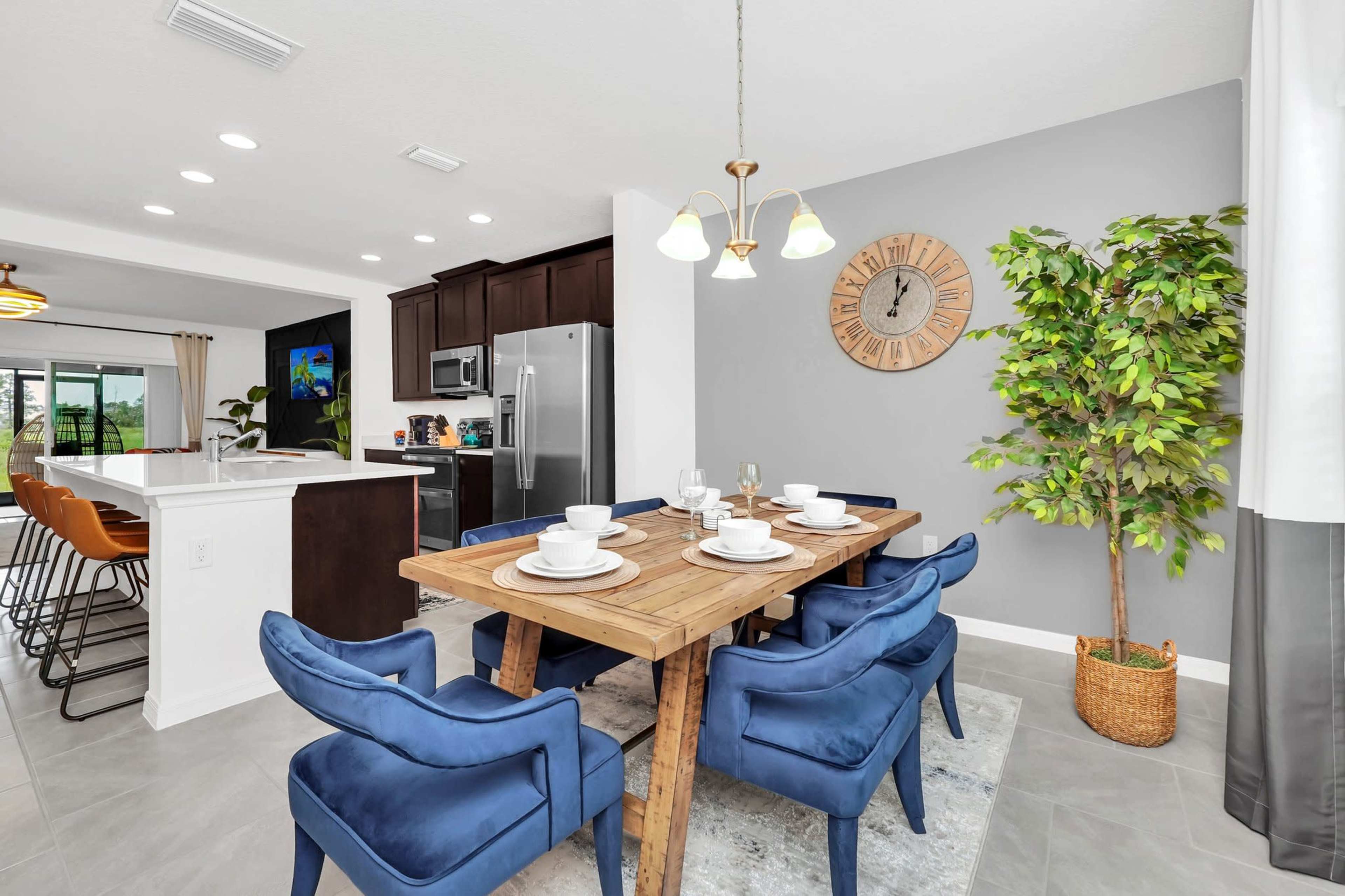 A dining area features a wooden table set for six, surrounded by blue chairs, with a clock and a potted plant on the wall behind.