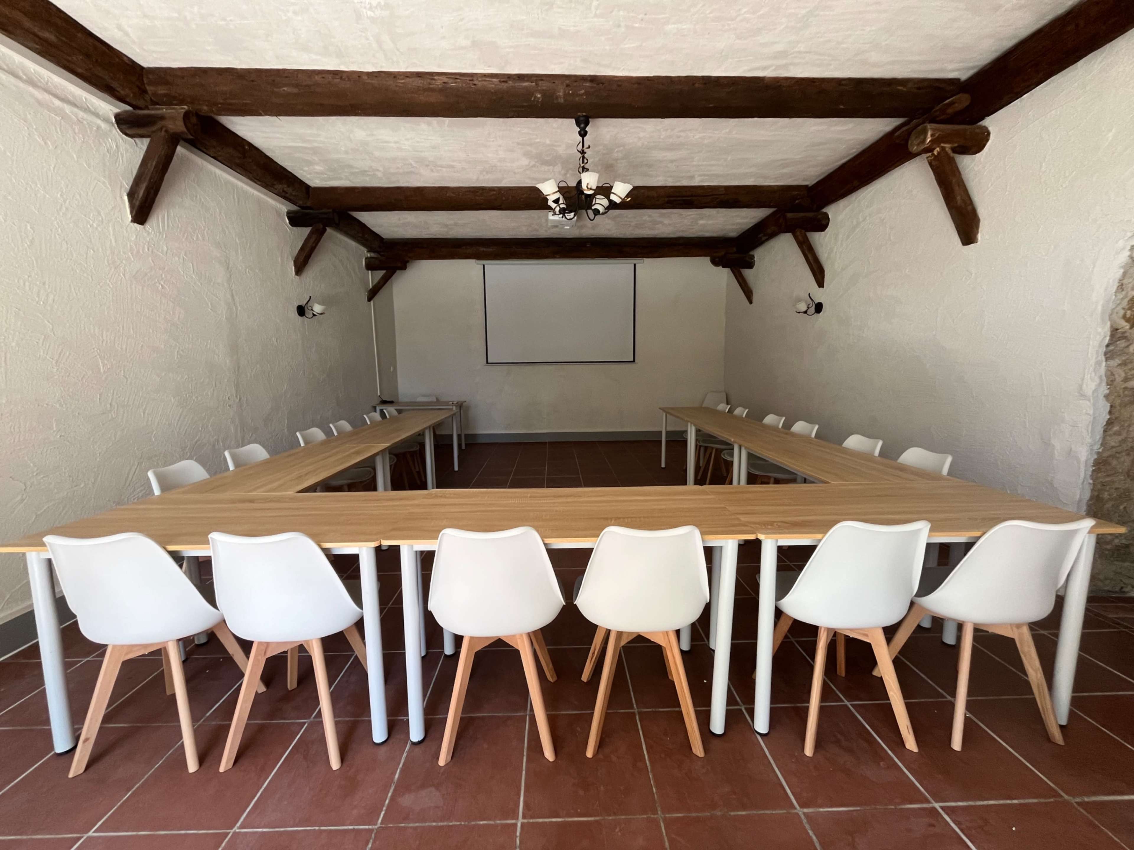 The image shows a conference room with a large, rectangular arrangement of tables and white chairs, all set against a textured wall and wooden beams.
