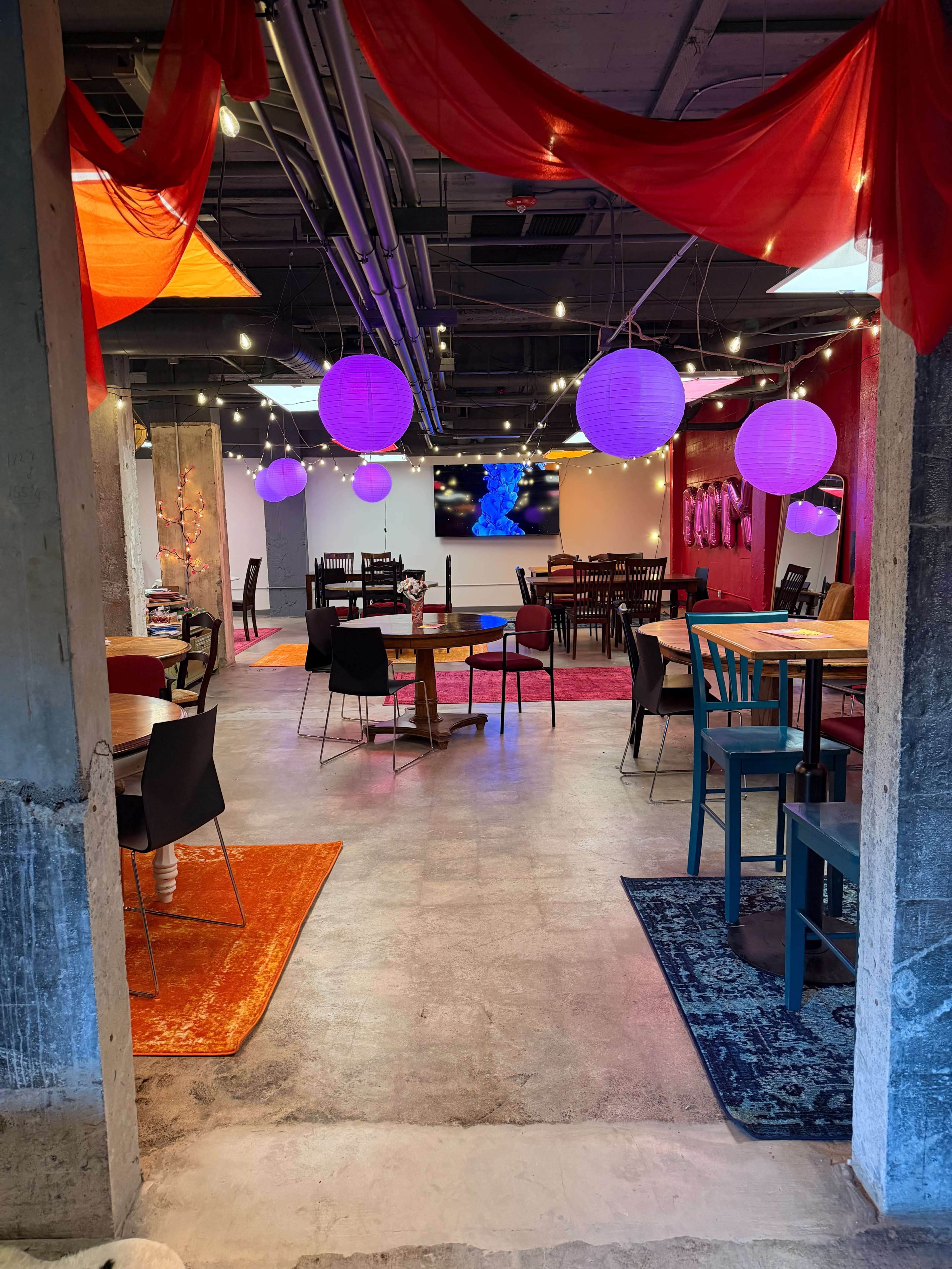 The image shows a modern indoor dining space with various types of tables and chairs, colorful lanterns hanging from the ceiling, and vibrant rugs on the floor.