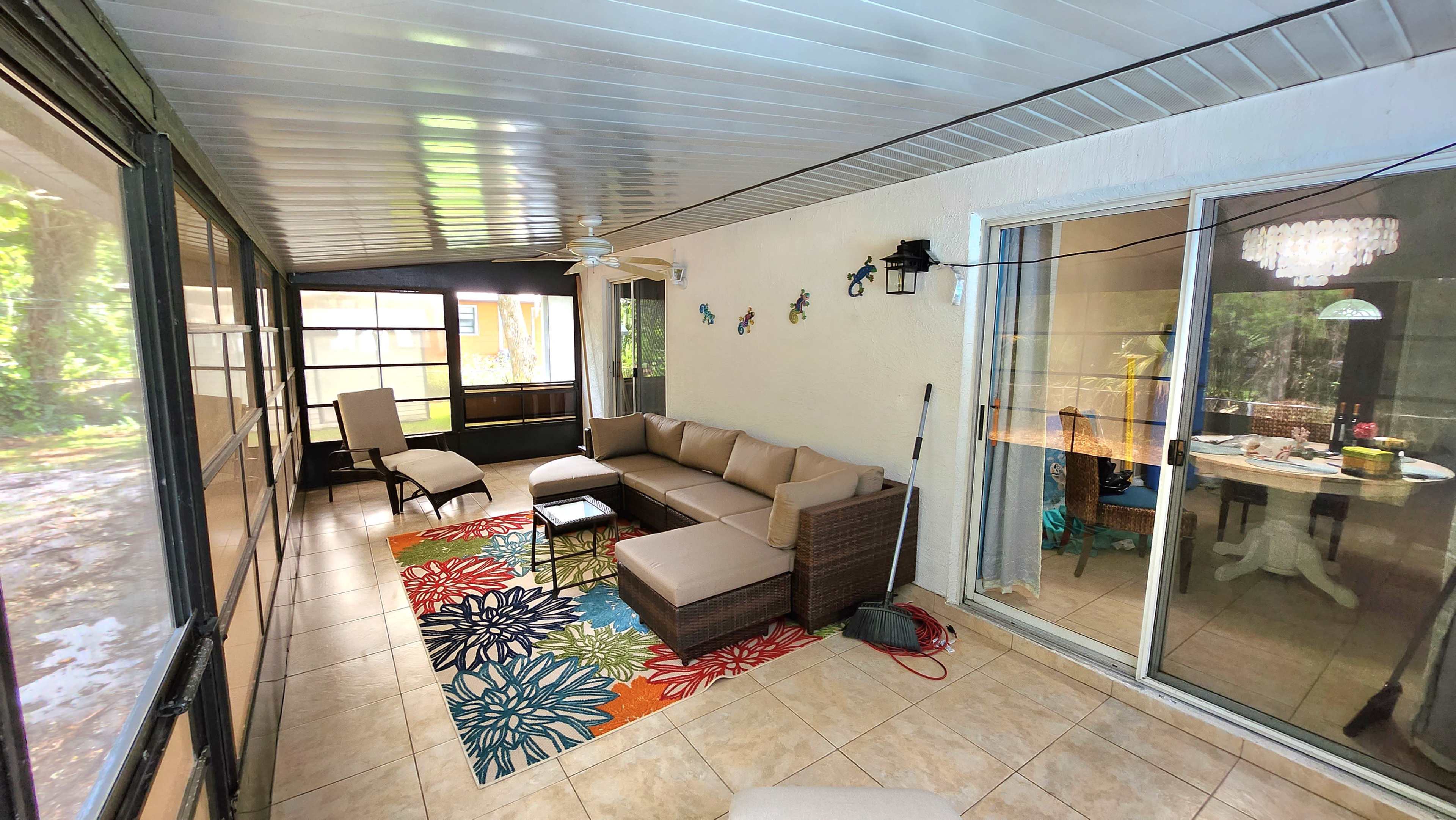 A sunroom with a seating area featuring a sectional sofa and coffee table, decorated with a colorful rug, and a dining area visible through sliding glass doors.