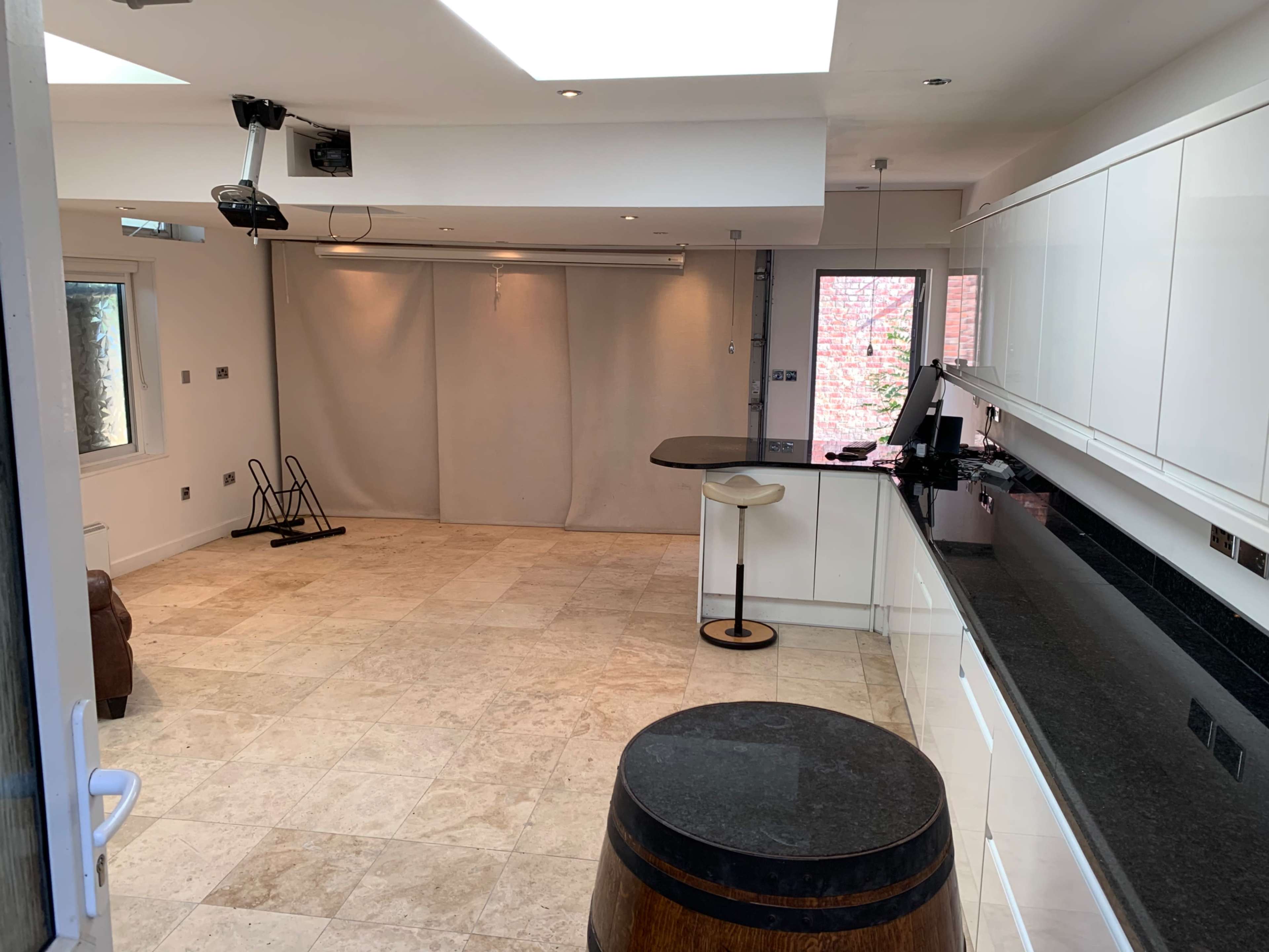 A spacious, neutral-toned room with a kitchen area, a bar stool, a projector mounted on the ceiling, and a large, blank wall for projection.