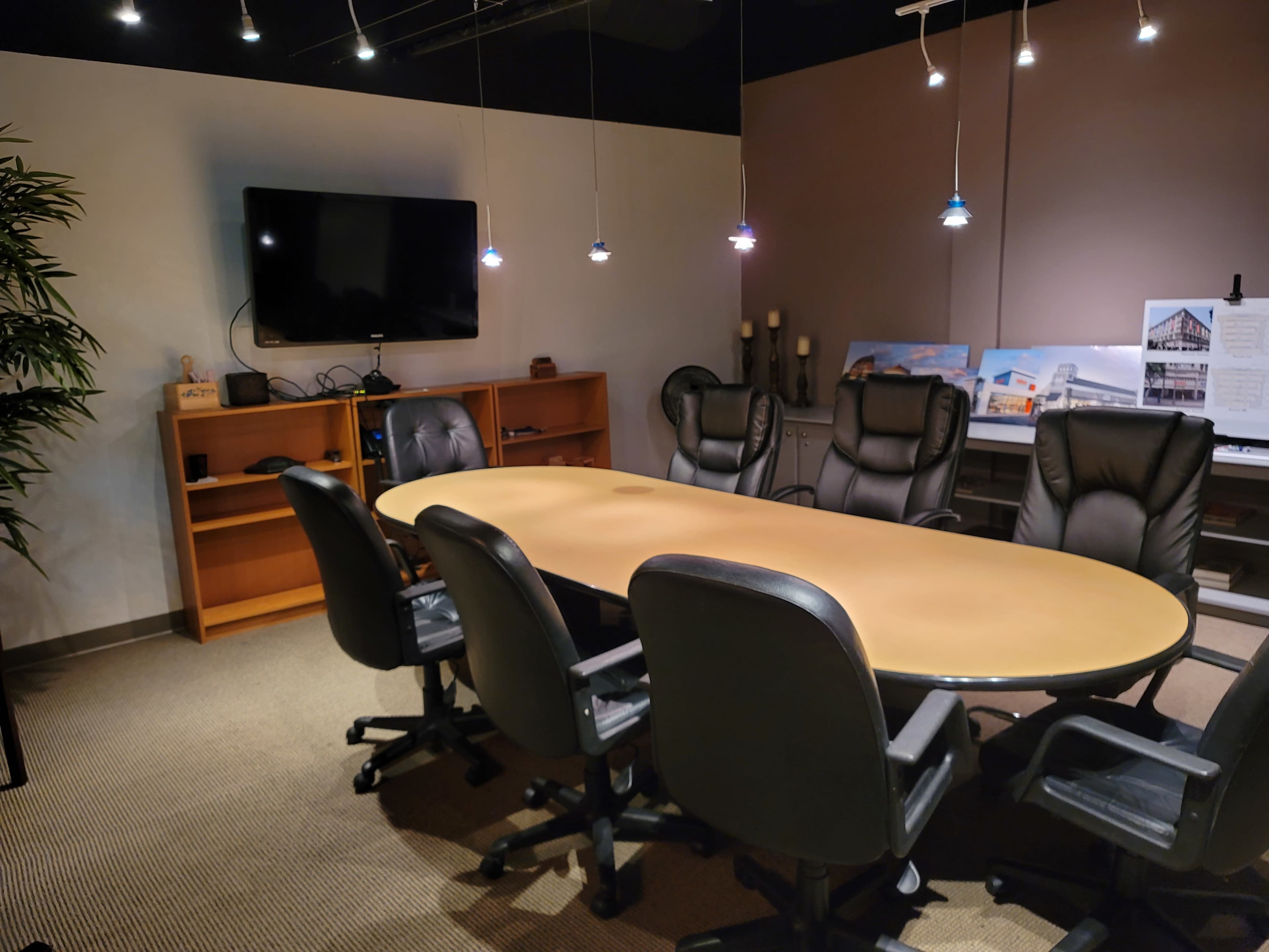 A modern conference room featuring a large oval table surrounded by six black chairs, with a television mounted on the wall and shelves containing displays and lighting fixtures.