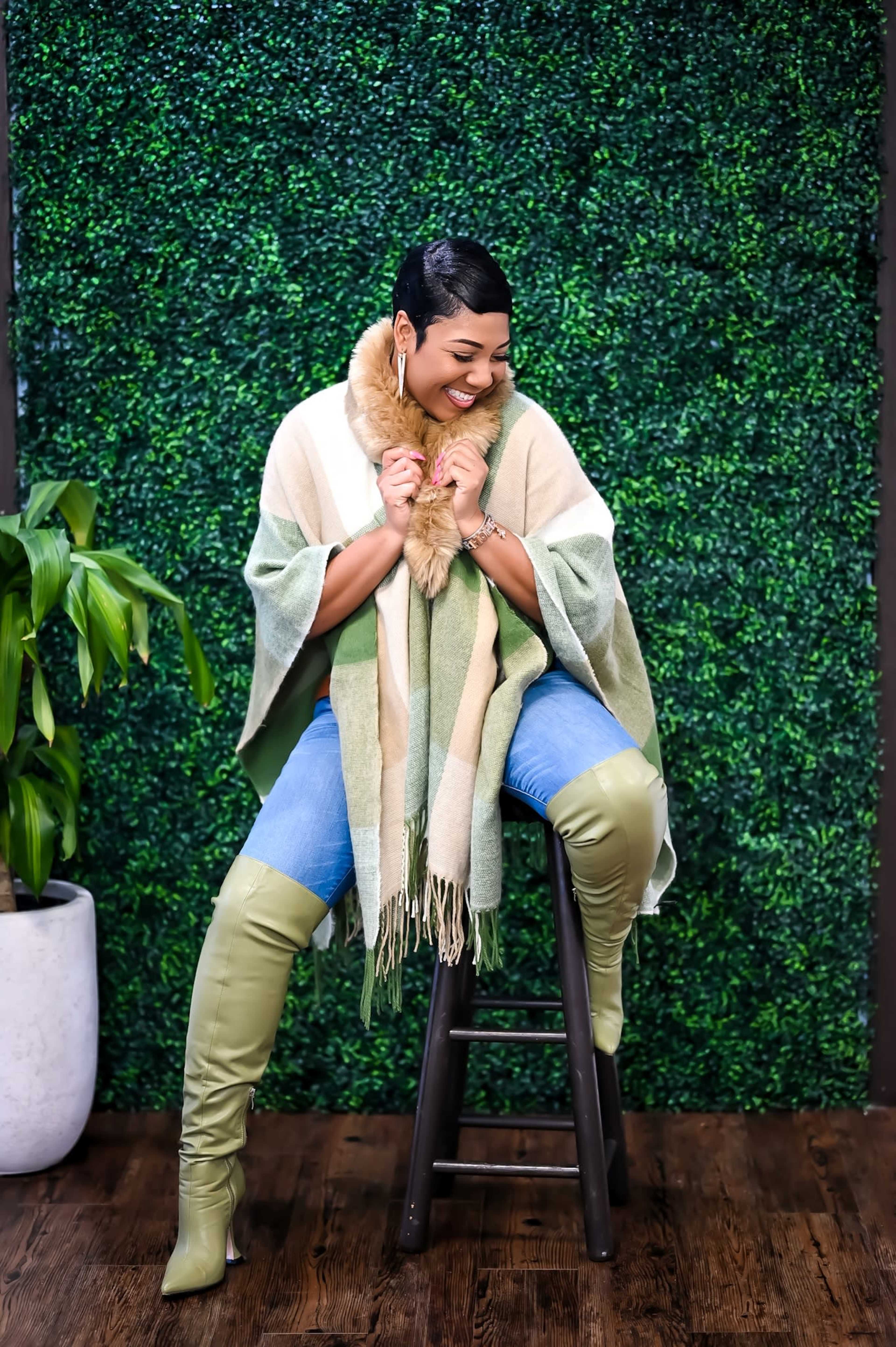 A person sits on a wooden stool, wearing a green and beige poncho with a fur collar, paired with tall green boots, against a backdrop of artificial greenery.