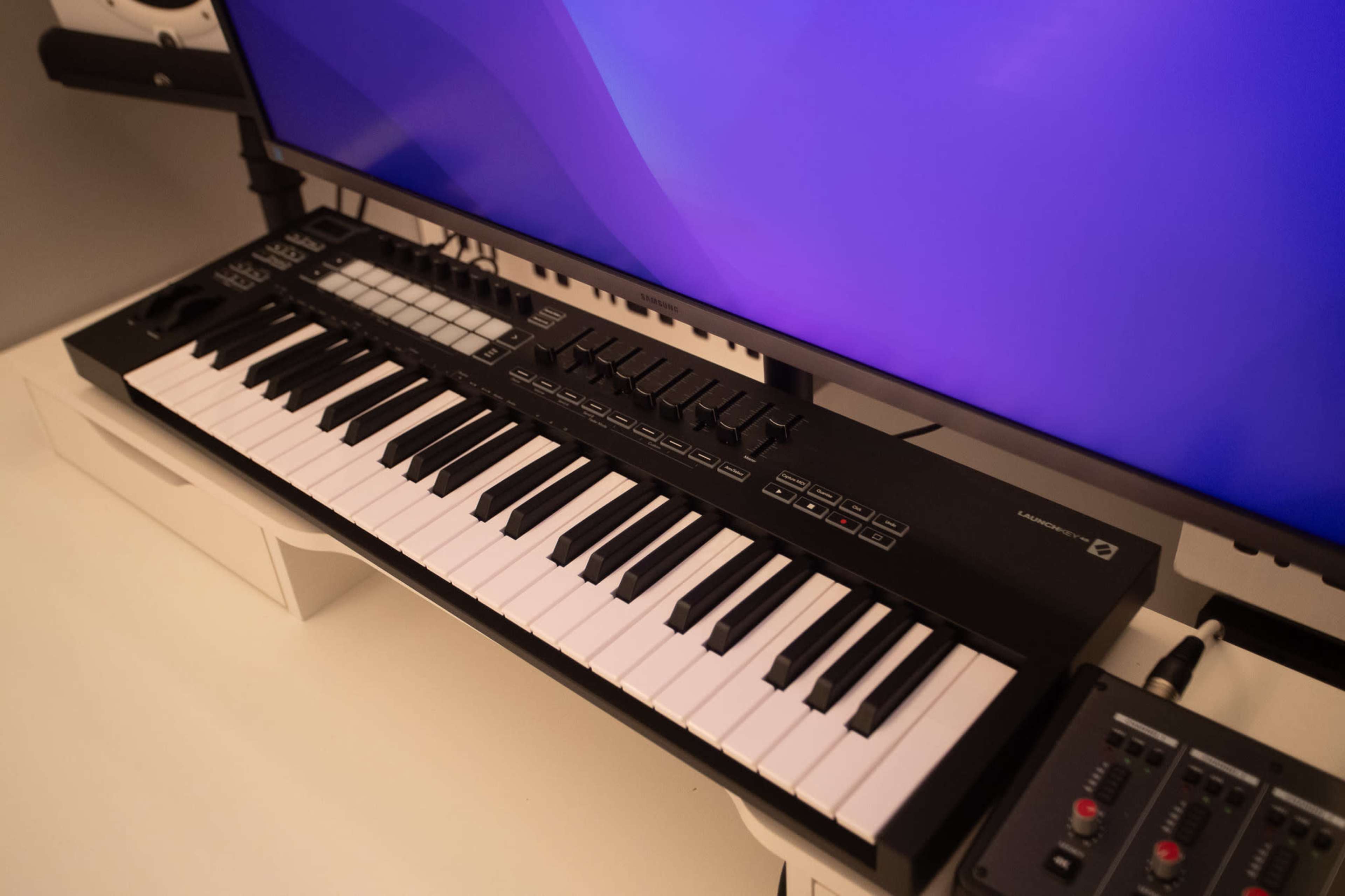 A MIDI keyboard is positioned on a desk in front of a computer monitor displaying a purple screen.