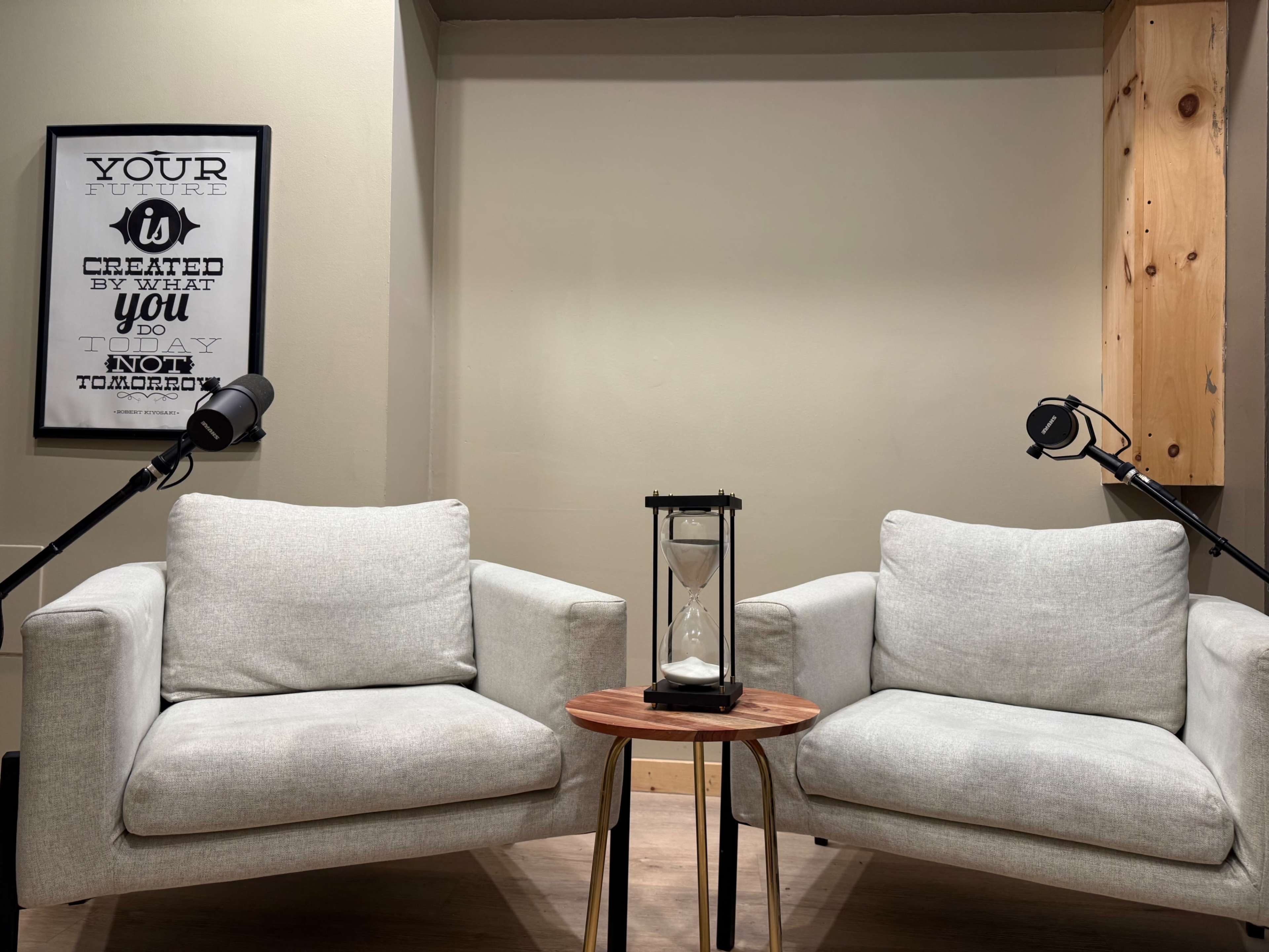 The image depicts two light gray armchairs facing each other, separated by a wooden side table with an hourglass, under a wall art piece that has motivational text.