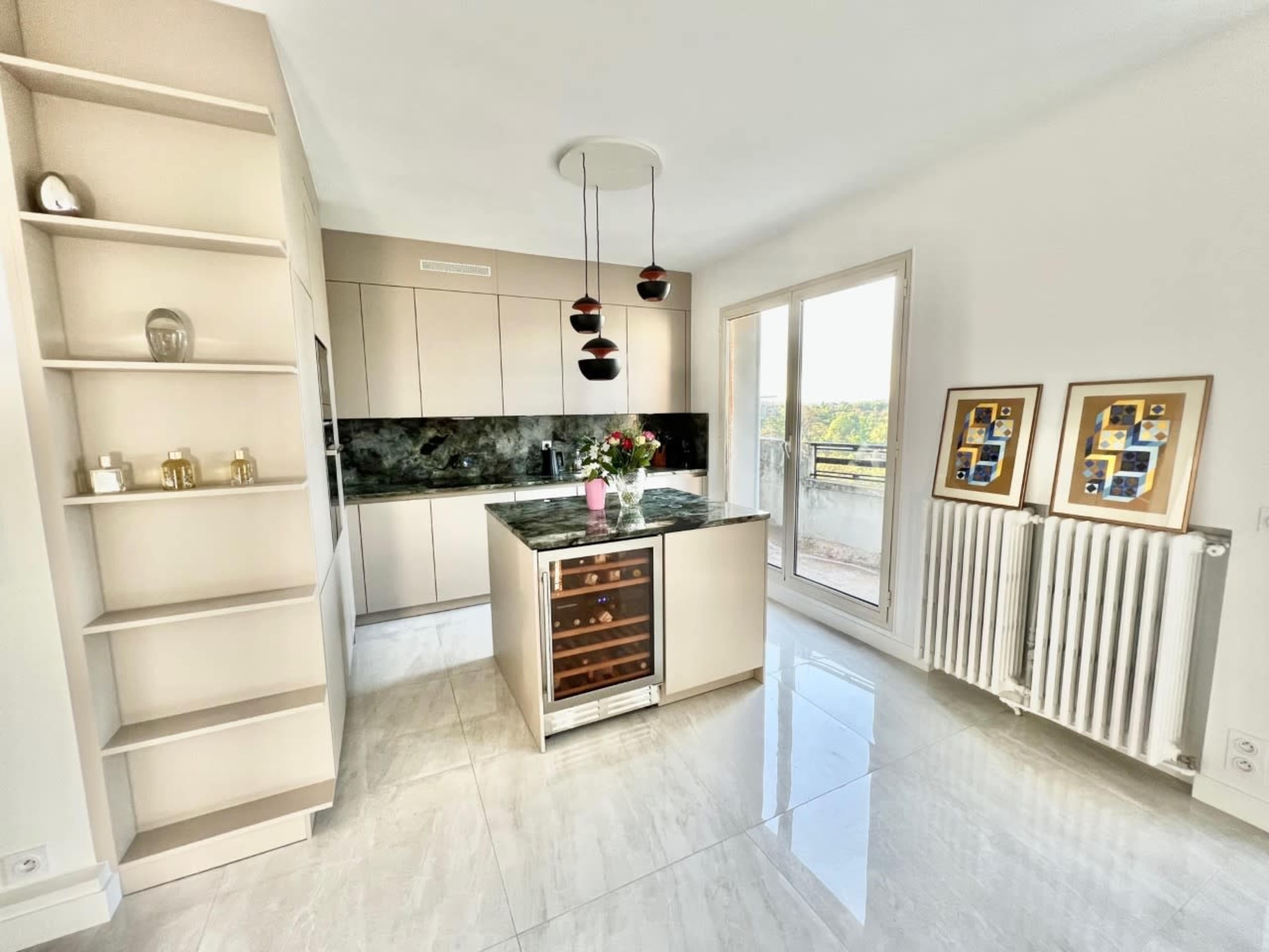 A modern kitchen with beige cabinets, a dark countertop, a wine cooler, and large windows that provide natural light.