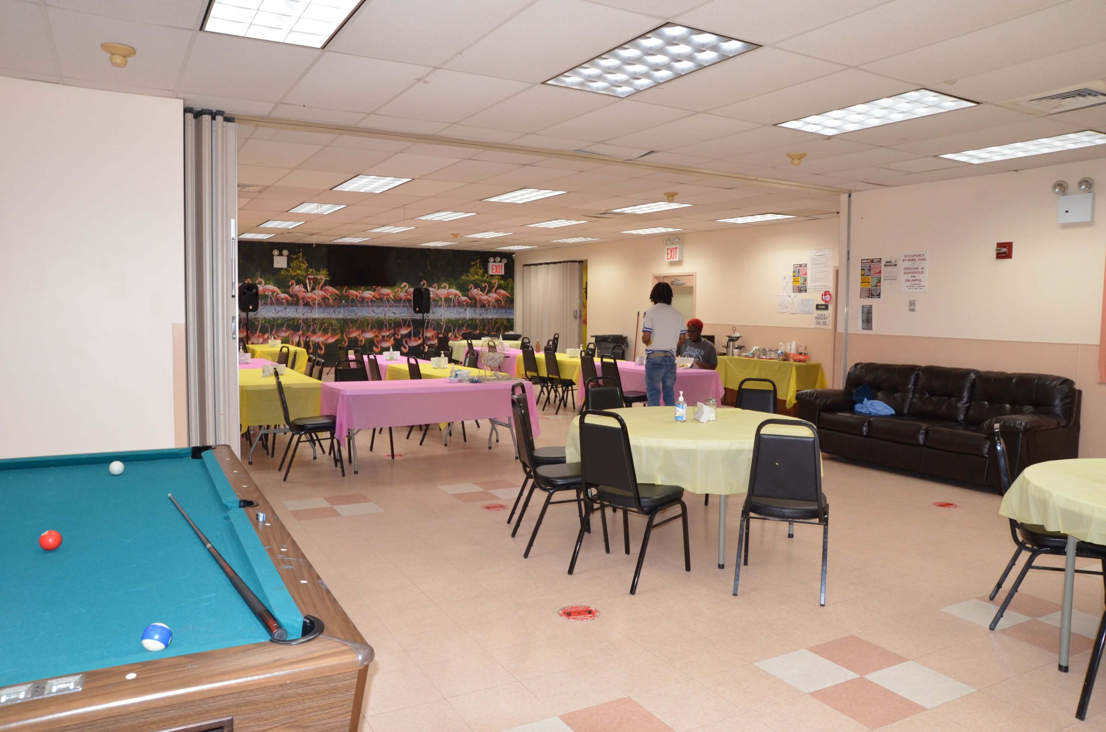 A spacious indoor room with several tables covered in yellow and pink tablecloths, a pool table on one side, and a couch along the wall.