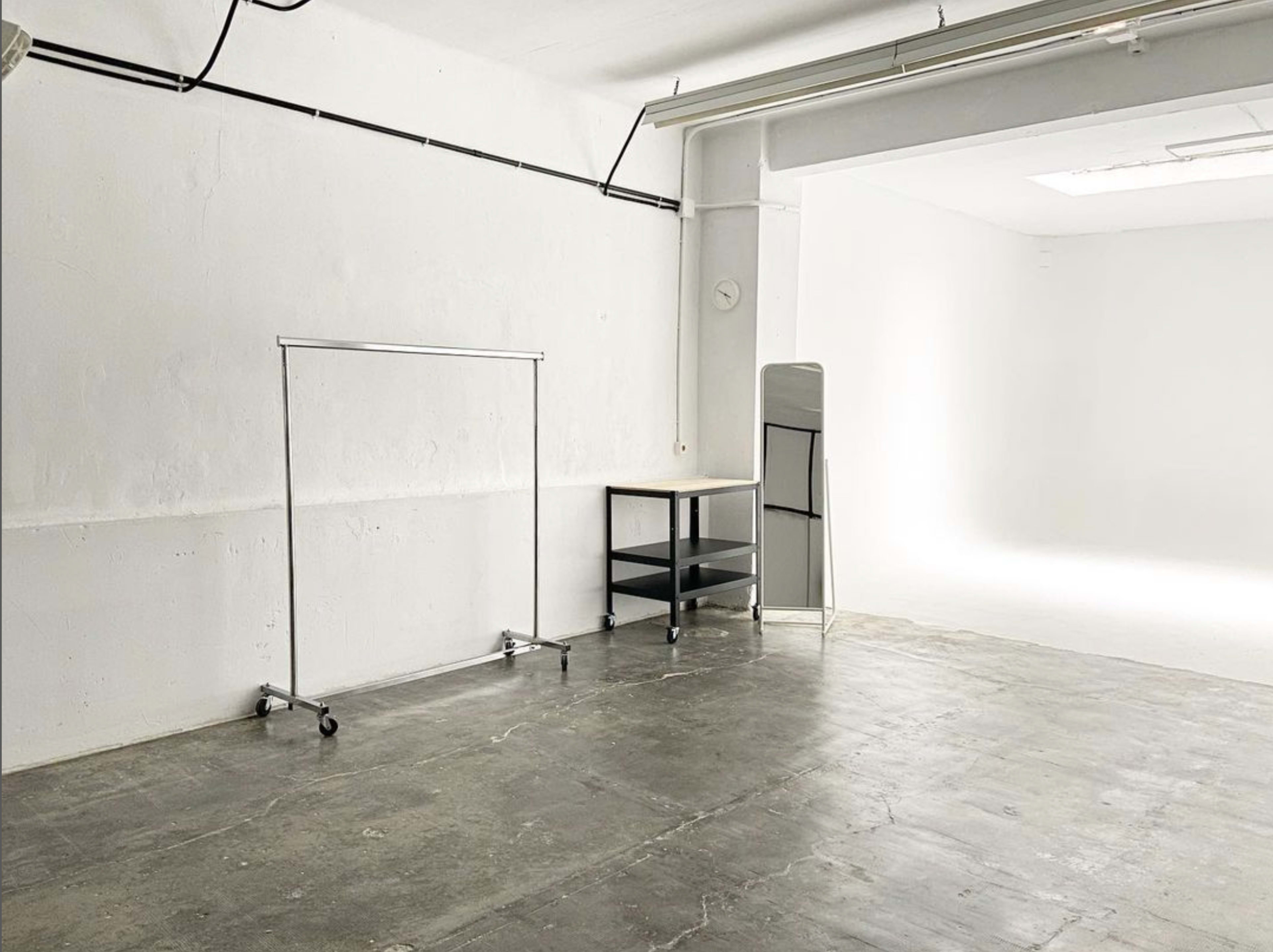 A minimalist, empty studio space with a clothing rack, a small shelving unit, and a mirror against a plain white wall.