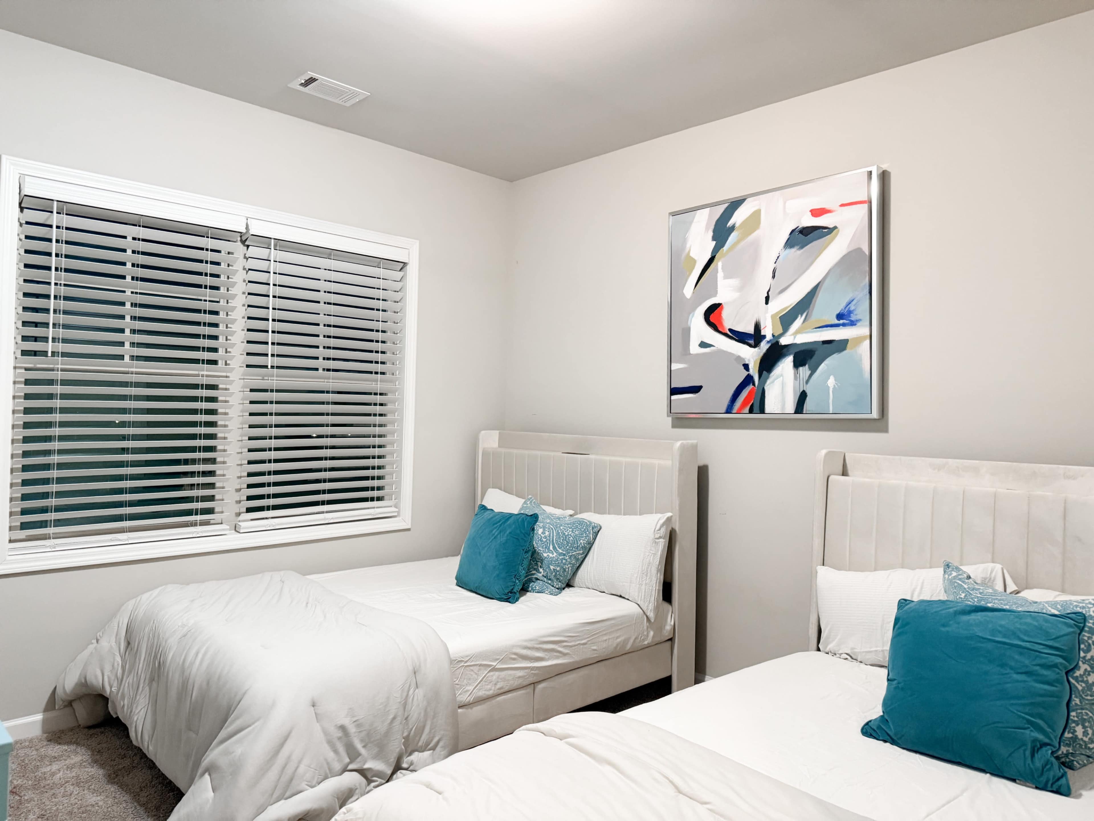 The image shows a bedroom with two twin beds, each adorned with white bedding and blue accent pillows, alongside a large abstract painting on the wall and a window with white blinds.