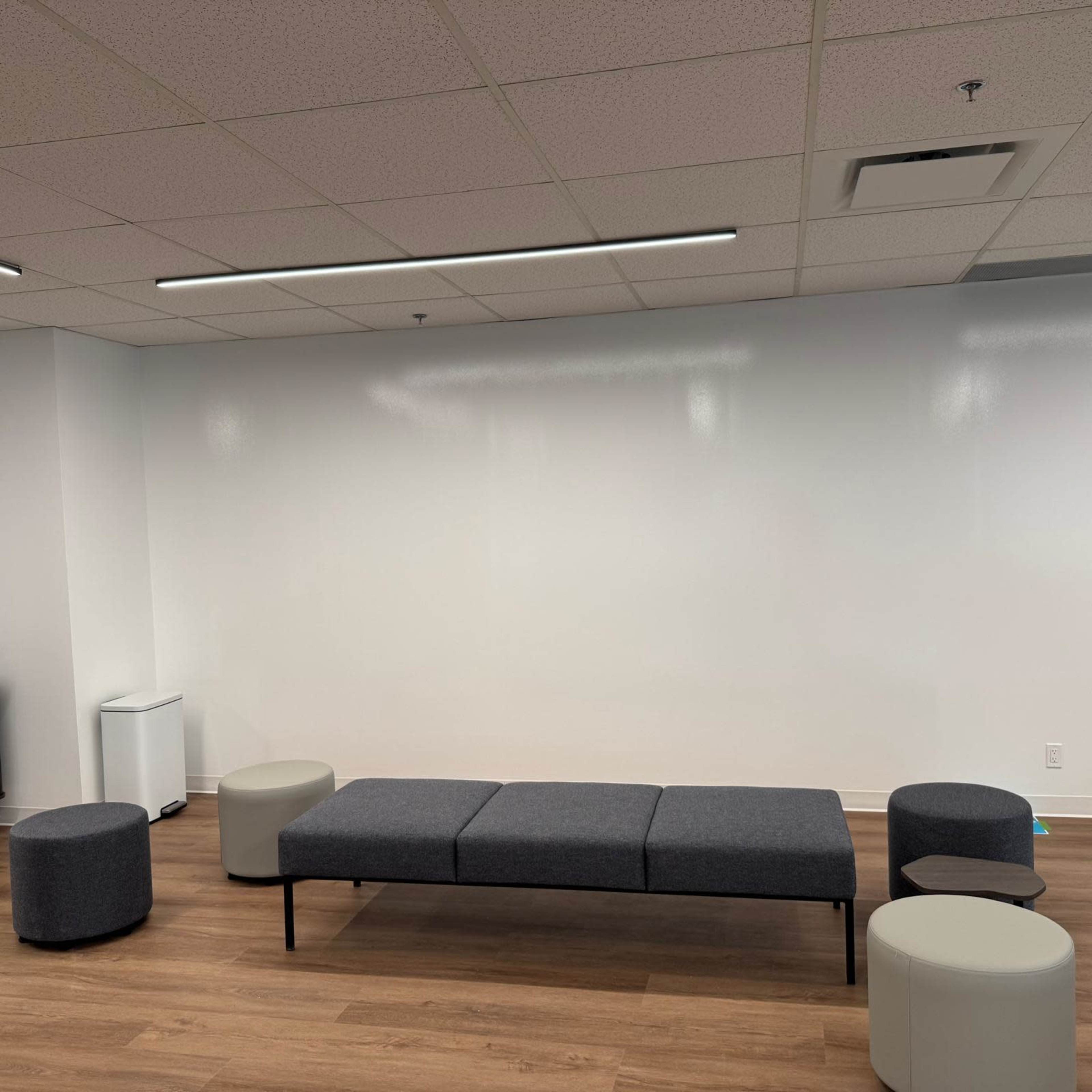 The image shows a modern, empty lounge area with a long gray bench and several round stools, set against a white wall.