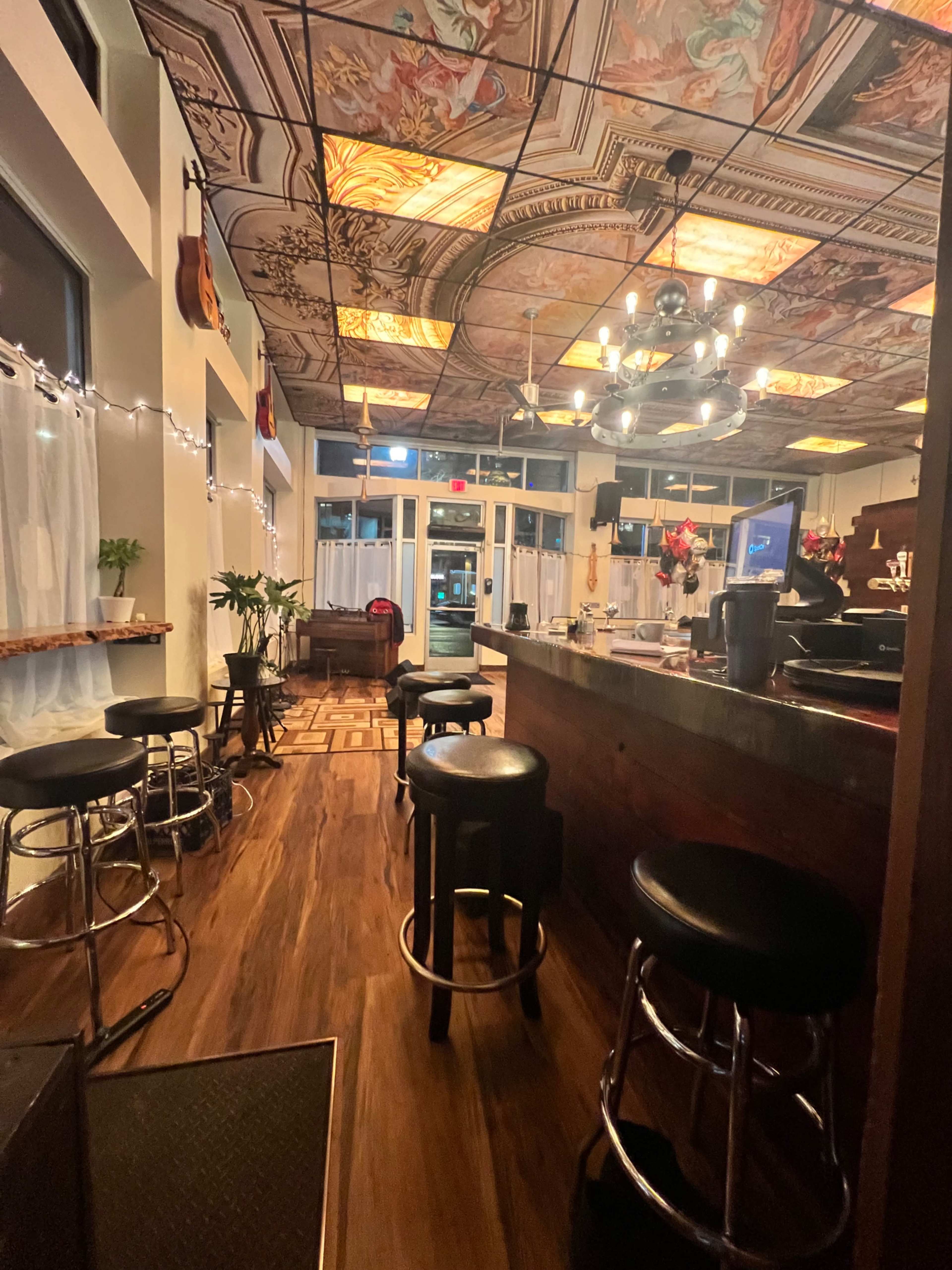 The image shows a dimly lit coffee shop interior featuring a bar with several stools, decorative ceiling tiles, and large windows that open to the street.