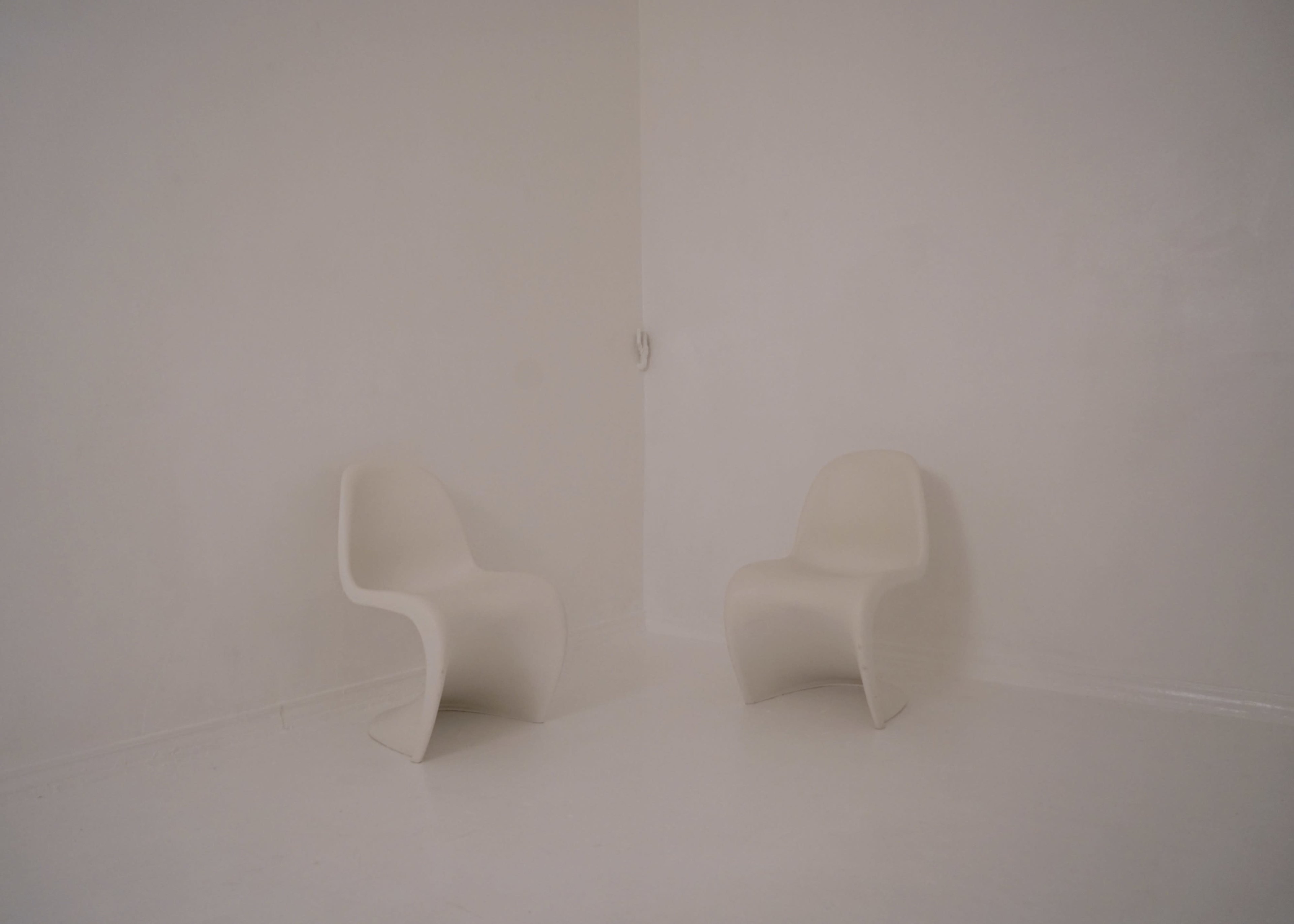 Two minimalist chairs are positioned facing each other in a stark, white corner room.